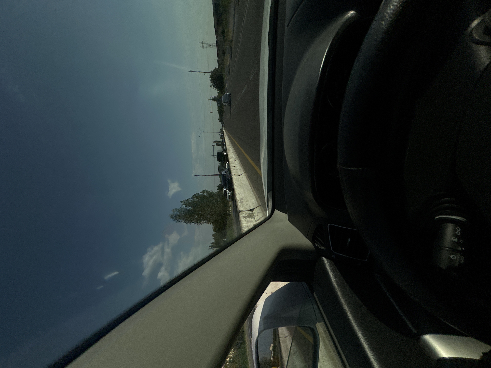The image depicts a view from the driver's seat of a car, looking out onto a road. The perspective is tilted, suggesting the photograph may have been taken while the car was in motion or the camera was held at an angle. The scene is set during the daytime, with a clear sky and scattered clouds, indicating fair weather.
The primary objects are the car's interior, including the steering wheel and dashboard components, and the road outside. The road appears to be a multi-lane highway or a major road with a yellow line marking the center. There are other vehicles visible on the road and parked along the side, suggesting moderate traffic. Trees and vegetation line the roadside, with some power lines and poles also present in the distance. A power pylon is visible on the horizon, a common sight in rural or semi-rural areas. The overall setting appears to be an open, possibly arid or semi-arid landscape, consistent with the provided location context of Tashtugizak, Uzbekistan.
No people are clearly visible within the frame. The activity taking place is likely driving, or observing traffic from a stationary or slow-moving vehicle. Notable details include the reflection in the car's side mirror, showing a glimpse of the sky and roadside scenery. The lighting suggests it is daytime, possibly late morning or afternoon. There is no visible text within the image that provides specific information. The overall mood is neutral, focusing on the observational aspect of the road and environment.