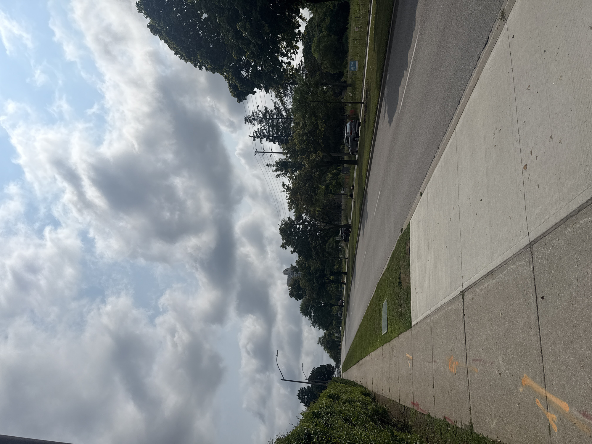 The image depicts a street scene in the city of Richmond Hill, Canada, on a cloudy day. A concrete sidewalk runs along the left side of the frame, with orange markings visible on some of its sections. Adjacent to the sidewalk is a strip of green grass, followed by a two-lane asphalt road. Several cars are visible driving or parked on the road and along its shoulder. Mature trees line the right side of the road, and in the distance, a multi-story building can be seen. The sky is filled with large, grey clouds, with patches of blue sky peeking through.