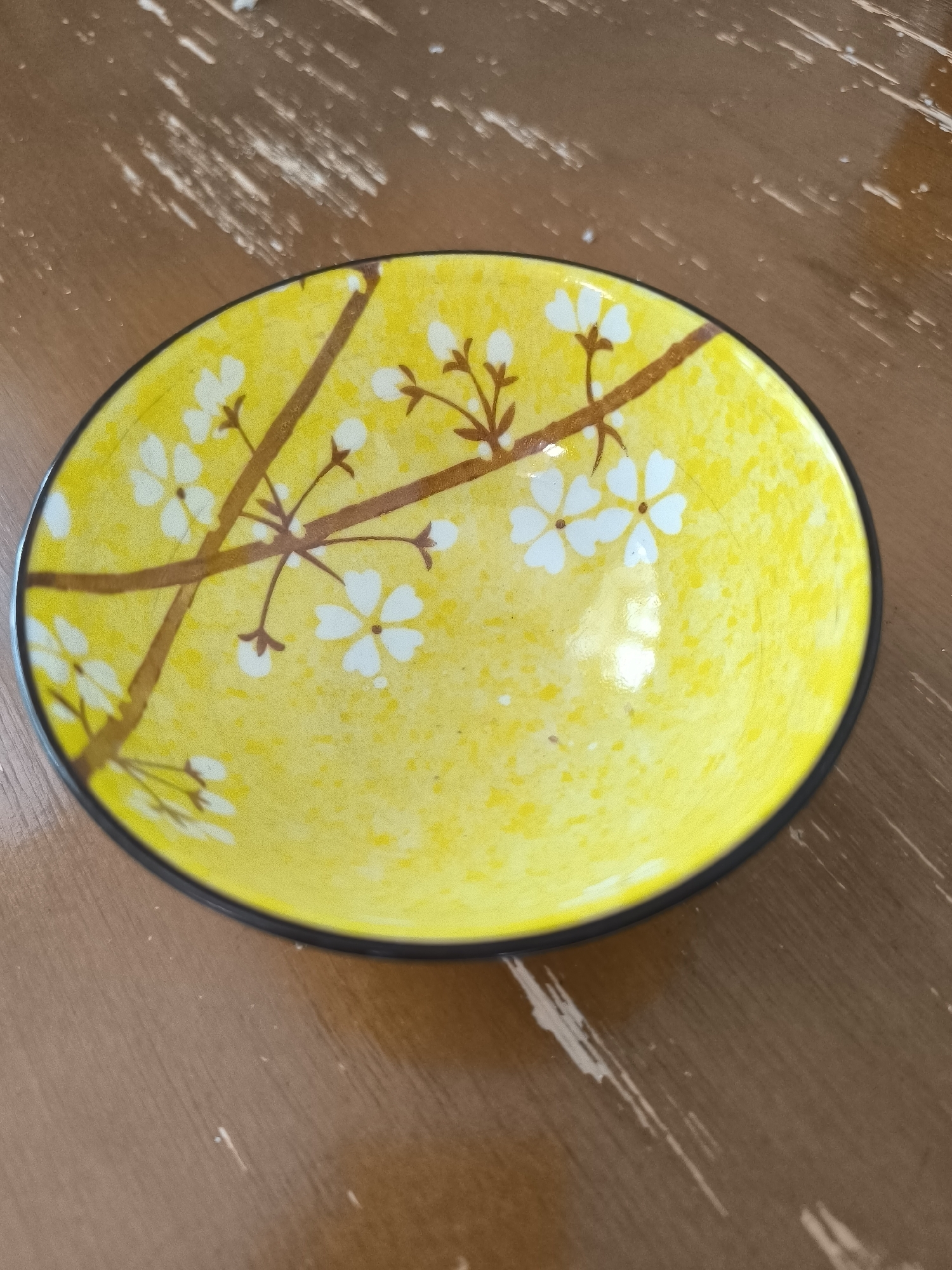 A yellow ceramic bowl with a cherry blossom design sits on a wooden table. The bowl has a black rim and the interior features brown branches and white blossoms. The table surface is brown and shows a distressed wood grain with white streaks. The scene is indoors. No location in Oskemen, Kazakhstan, can be determined from the image.