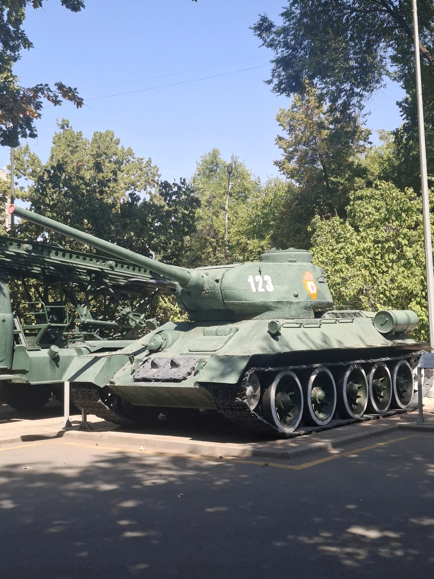 A military tank, painted olive green, is parked outdoors on a paved surface. The tank is oriented facing the right side of the frame. It has a long barrel extending forward. Visible markings on the tank's turret include the number "123" in white and a red and yellow emblem. To the left of the tank, part of a large, green, metal structure with a ladder is visible. The background consists of lush green trees and a clear blue sky. The ground in front of the tank is paved, with yellow lines marking a parking area and shadows from trees cast upon it. A thin wire is visible against the sky in the upper portion of the image. The scene appears to be a static display, possibly in a park or memorial site, located in Almaty, Kazakhstan, given the presence of T-34 tanks which are often displayed in Soviet-era war memorials.