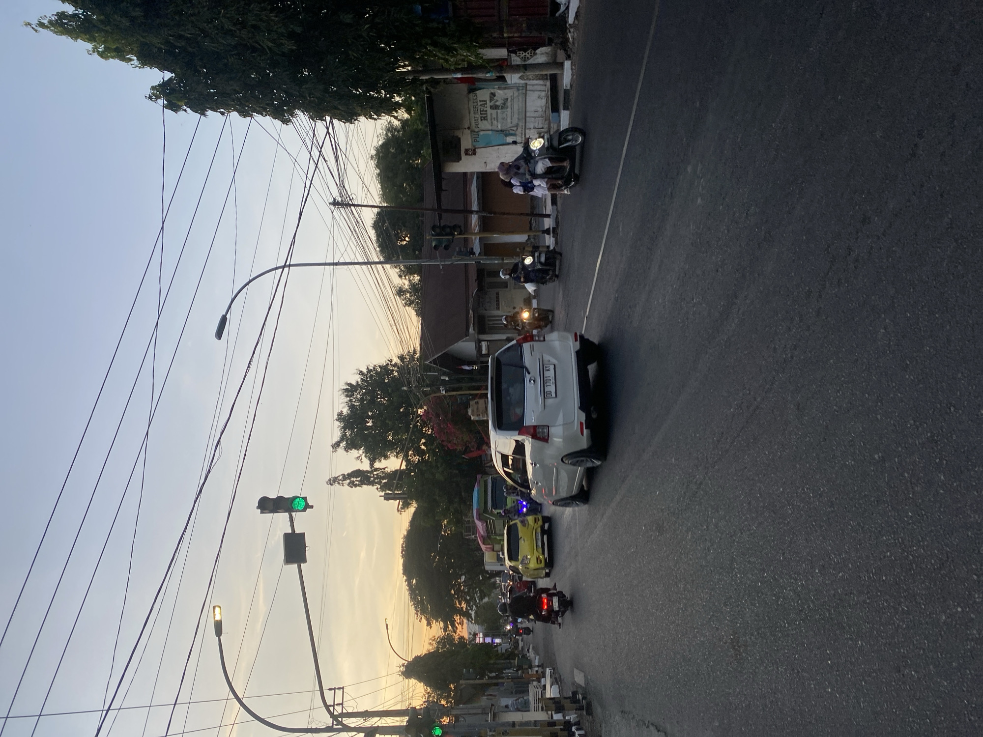 The image depicts a street scene in Bantaeng, Indonesia, at dusk. A white car with license plate DD 1701 KY is in the foreground, moving on the right side of the road. Several motorcycles and a yellow car are also present in traffic. Traffic lights are visible and appear to be green. The sky is illuminated by the setting sun, casting a warm glow. Trees and buildings line the street, and numerous overhead wires crisscross the sky. The road is asphalt and appears to be in good condition. A few pedestrians are visible on the sidewalk in the distance.