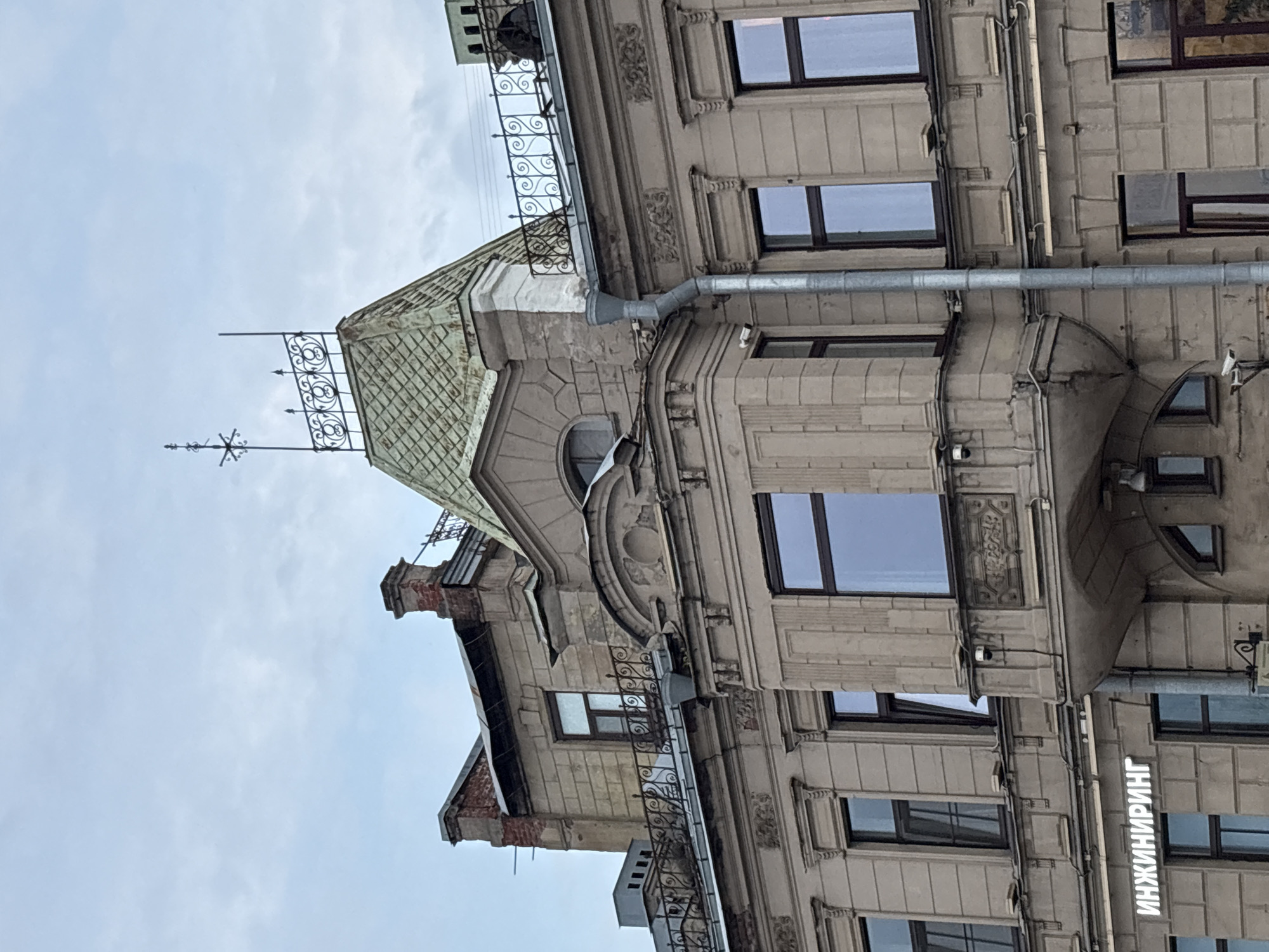 The image shows a close-up view of the exterior of an ornate, old building, likely in Saint Petersburg, Russia, given the visible Cyrillic text. The architecture suggests an early 20th-century style, with intricate stonework, decorative elements, and a mansard roof. Several windows with dark frames are visible, some reflecting the overcast sky. A metal downspout runs along the facade. A distinctive feature is a green, diamond-patterned mansard roof with a decorative metal railing and a weather vane-like structure on top. The building appears to be multi-story, with different architectural sections. A portion of the facade on the right side of the image has a sign with vertical white Cyrillic lettering that reads "ИНЖИНИРИНГ" (Engineering). The overall impression is of a historical urban setting on an overcast day, with the architecture being the primary focus. There are no people visible, and the scene is static. The weather appears to be cloudy, contributing to the muted colors of the building and the sky.