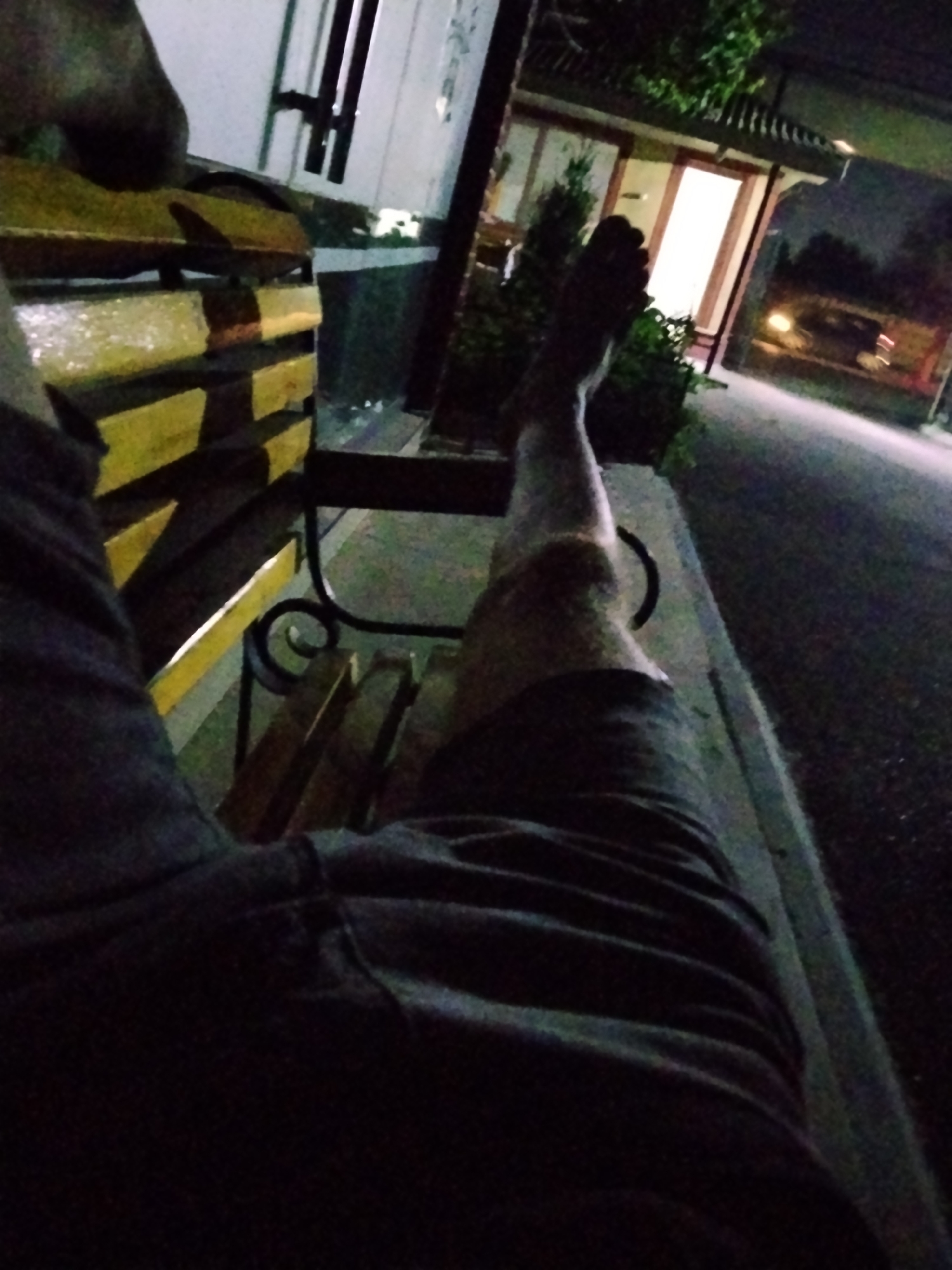 The media file captures a first-person perspective of a person resting on a bench at night. The scene is dimly lit, suggesting it is late evening or night. The subject's legs are visible, one extended and bare, with the other partially obscured by dark clothing. The bench appears to be made of wood with yellow markings and is set outdoors. Behind the bench, a building with illuminated windows and a traditional-style roof can be seen. Adjacent to the building, there is a paved area, possibly a sidewalk or road, with faint headlights from a distant vehicle. Green foliage is also visible, indicating an outdoor setting, possibly a park or a commercial area with landscaping. The overall mood is relaxed and perhaps contemplative, given the static posture and the quiet nighttime environment. There is no visible text in the image. The location context provided is Tashkent, Uzbekistan, which helps to frame the setting.