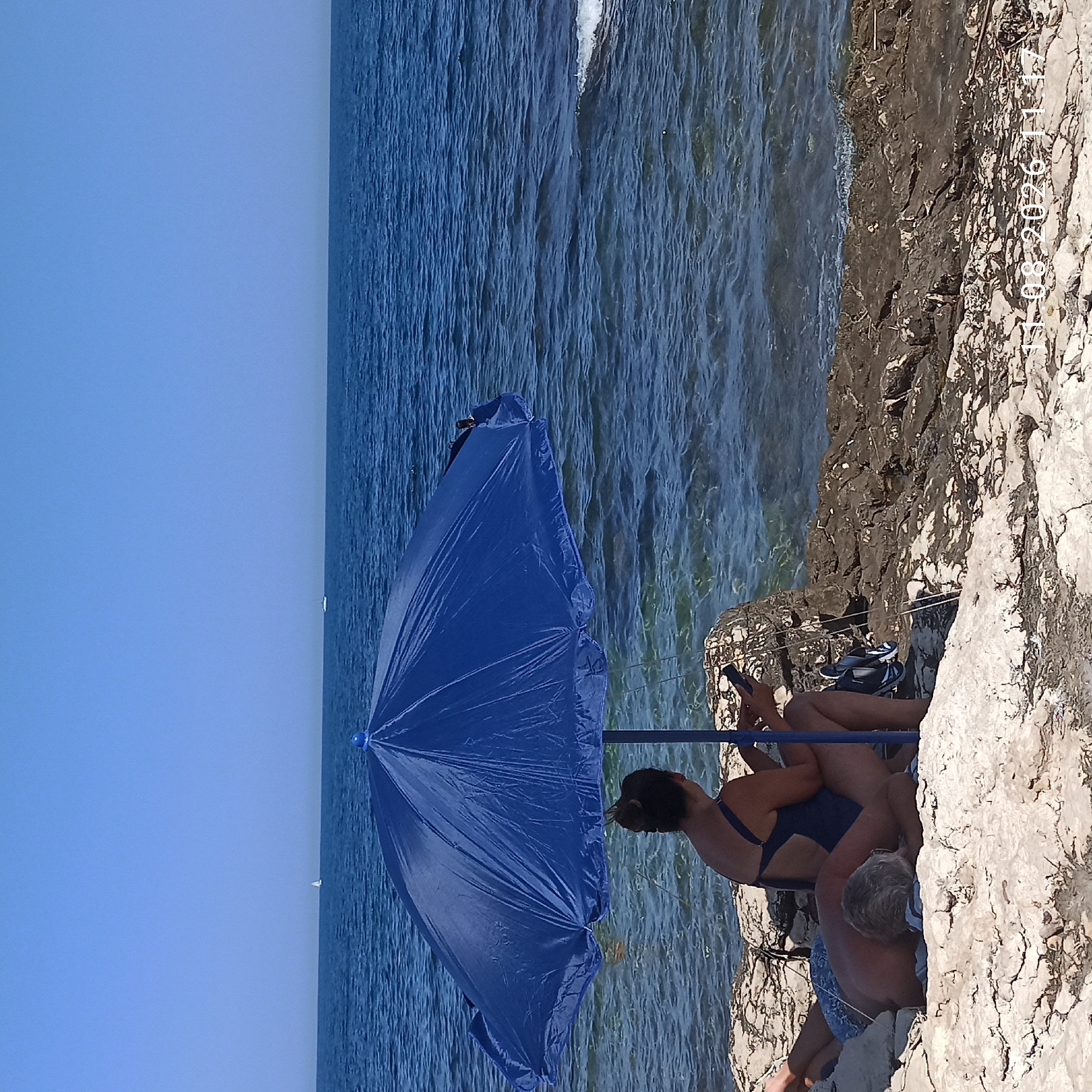A man and a woman are sitting on a rocky beach near the sea. The man is lying down on a towel, facing away from the camera, while the woman is sitting up, facing the sea and holding a mobile phone. They are both partially shaded by a large blue umbrella. The sea is calm and blue, with a clear horizon under a bright sky. The shoreline is made up of light-colored rocks interspersed with darker, rougher stones. There are no discernible buildings or landmarks to identify the specific location within Molfetta, Italy. The date and time stamp "11.08.2026 11:17" is visible in the lower right corner of the image.