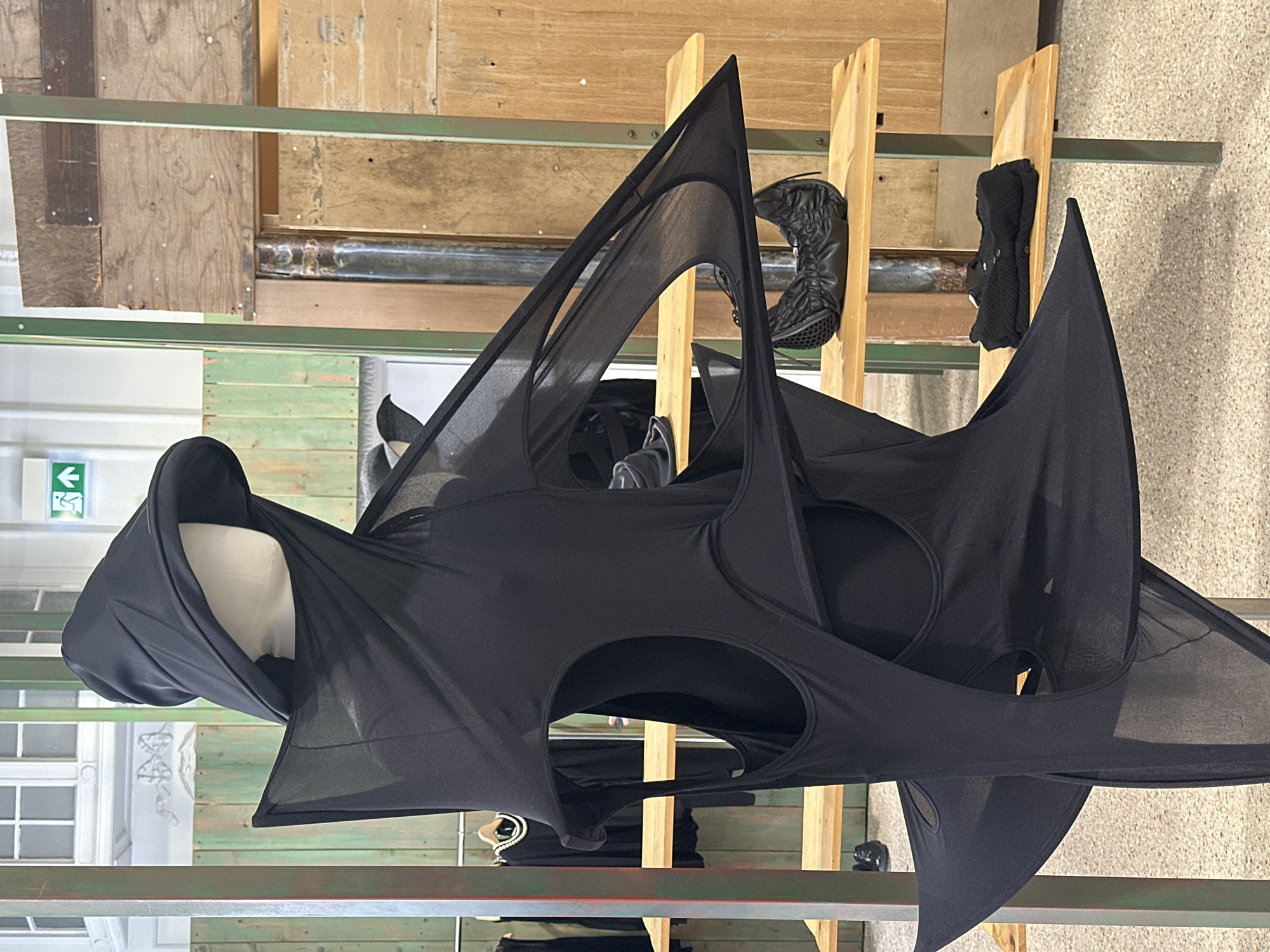 The image shows a black, geometrically-shaped, hooded garment displayed on a mannequin head on a clothing rack. The rack is made of green metal and appears to be in a retail or gallery space. Other clothing items and shoes are also displayed on the rack and on wooden structures nearby. The background includes a wall and window with a visible exit sign. The location is likely in London, United Kingdom, based on the architectural style and design details.