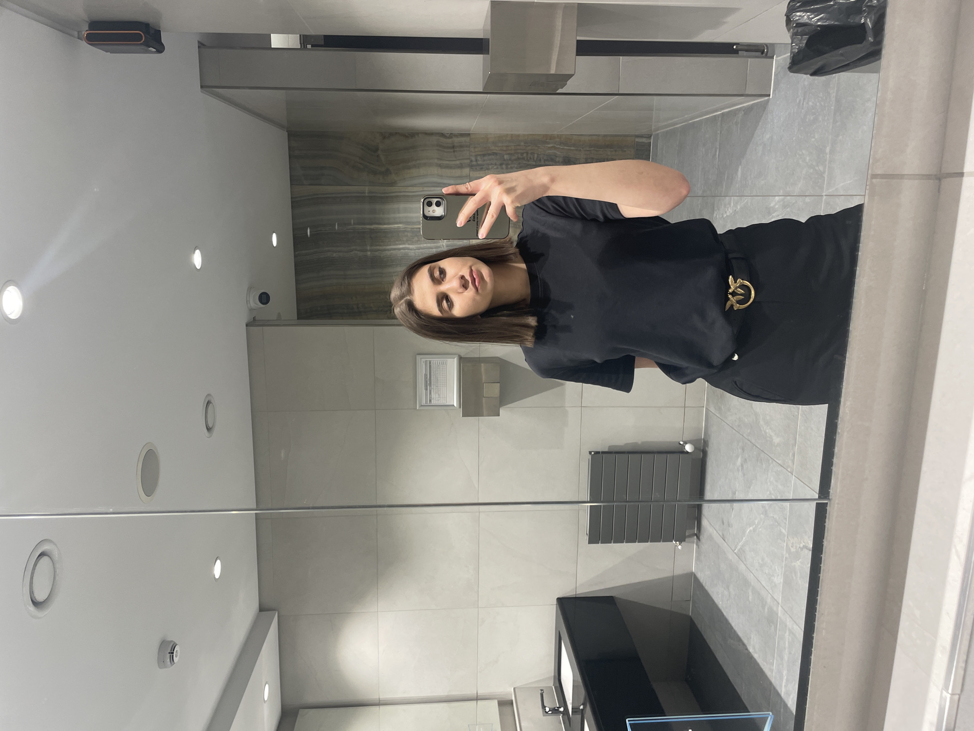 A woman is taking a selfie in a bathroom with a large mirror. She is holding a cell phone in her right hand and looking at the camera. The woman has medium-length brown hair and is wearing a black top and pants with a gold belt buckle. The bathroom has light grey tile walls and flooring. There is a black sink and counter beneath the mirror. A grey radiator is on the wall to the right of the sink. The ceiling is white with recessed lighting. The location is unknown.