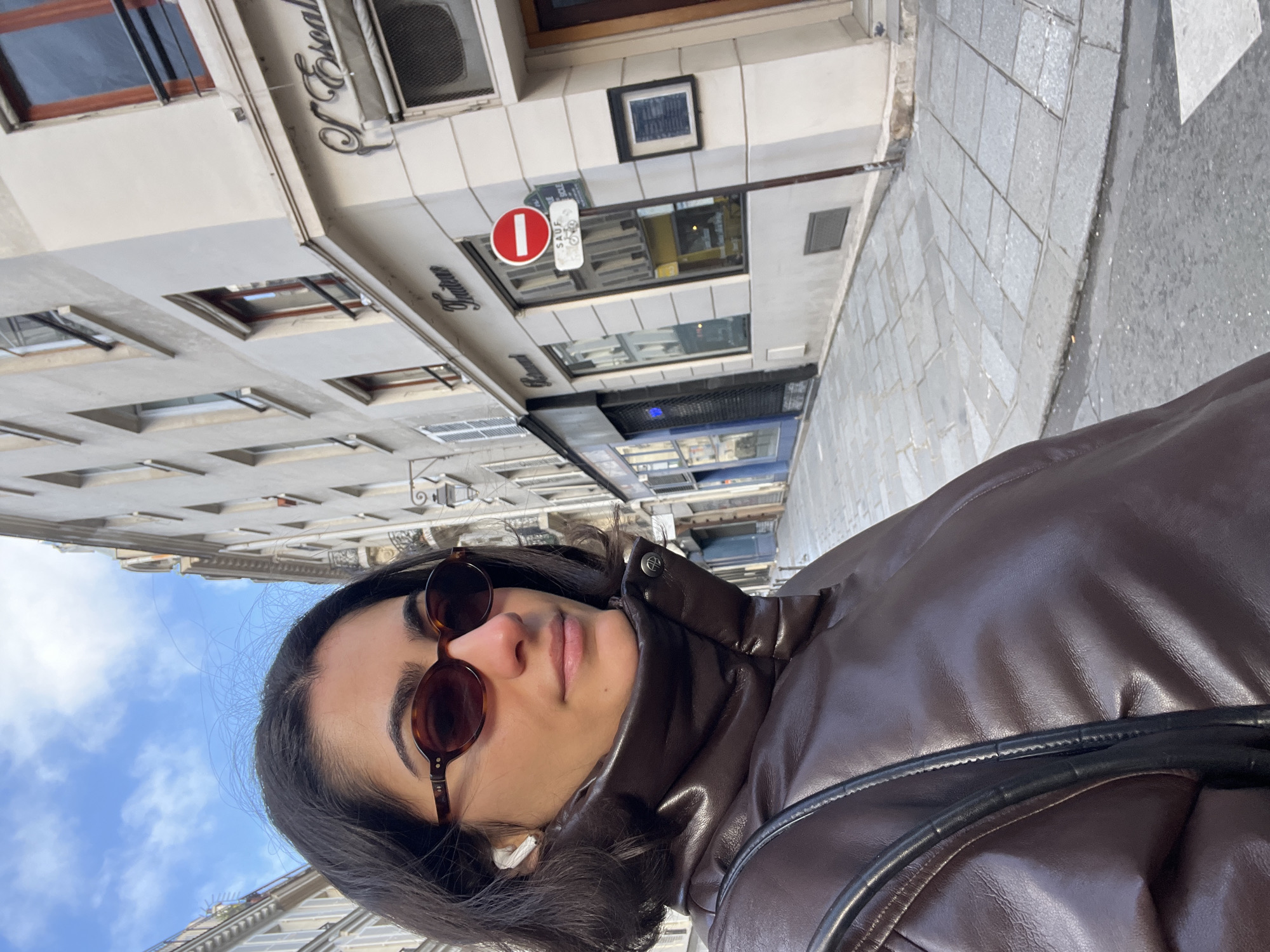 A street-level, angled perspective shot depicts a person wearing a brown leather jacket and round, brown sunglasses in Paris, France. The person is partially visible; their face is prominent, and a white earbud is visible in their left ear. A black bag strap crosses their shoulder. The background includes multi-story buildings with Parisian architecture under a blue sky with scattered clouds. A street sign with a red circle and a white horizontal line is present. The storefront "J. Esscal" is visible on one of the buildings. The sidewalk is paved with light-colored stone.