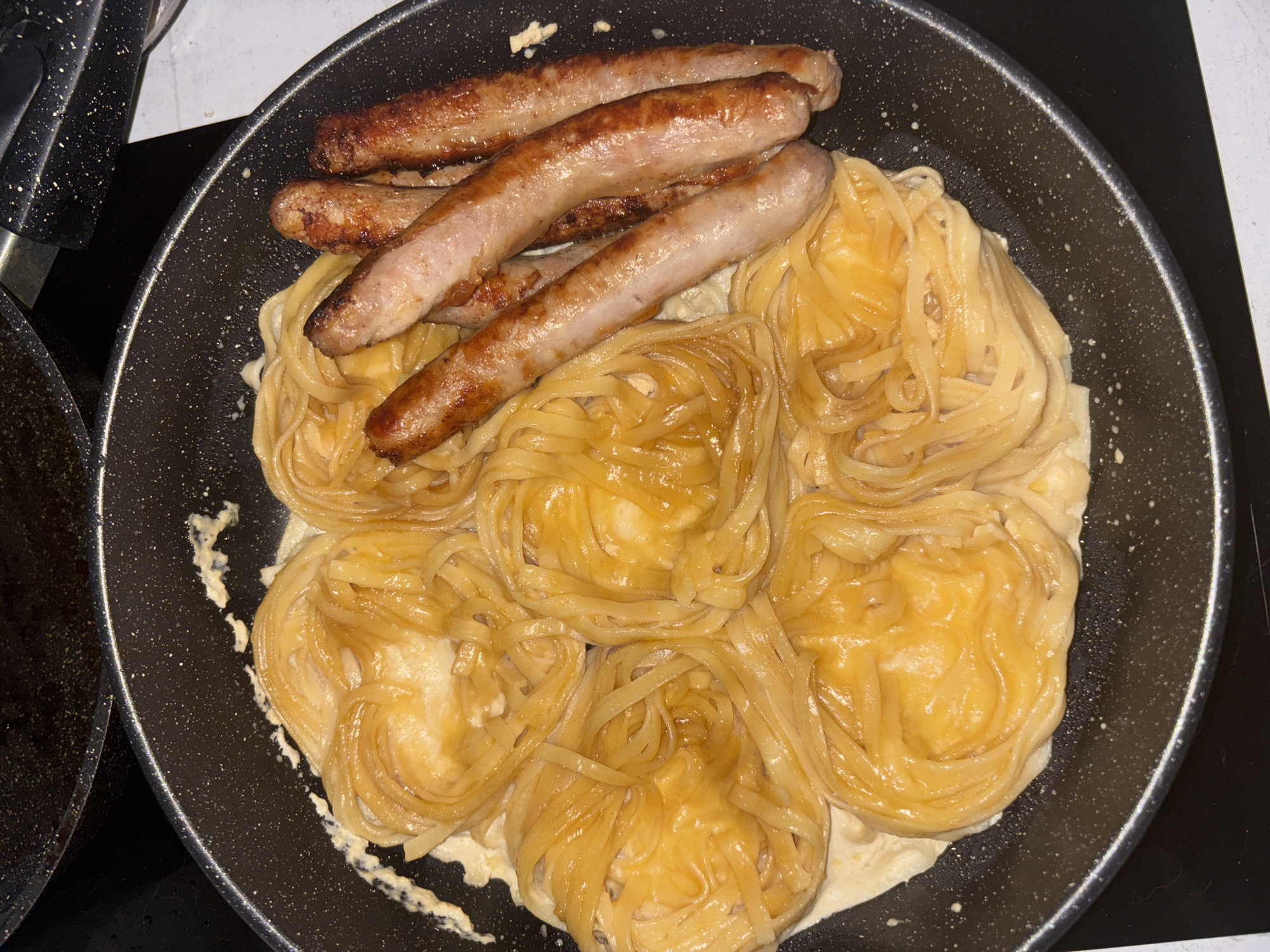A close-up, high-angle shot captures a frying pan containing a meal of sausages and pasta nests, likely prepared in a domestic kitchen in Vsevolozhsk, Russia.
The central subject is a black non-stick frying pan, featuring a speckled finish, occupying most of the frame. To the left, a portion of another similar pan with a black handle is partially visible. Inside the main pan, five browned sausages are arranged at the top, mostly lying diagonally. These sausages appear fully cooked, exhibiting a light to medium brown, slightly crisp skin with some darker, caramelized patches.
Below the sausages, and filling the majority of the pan, are six distinct nests of pasta. The pasta consists of flat, wide noodles, resembling tagliatelle or fettuccine, coiled into round, individual servings. The noodles are a warm, golden-yellow color and appear glossy, suggesting they are coated in a sauce or cooking liquid. Beneath and around these pasta nests, a light-colored, creamy sauce or liquid is visible, appearing to envelop the pasta. Some stray drops or splashes of this sauce are present on the pan's surface outside the main food area.
The background is simple, with part of a dark stovetop surface visible to the right and a lighter, possibly marble-patterned countertop or backsplash section visible behind the pan. The lighting is uniform, suggesting indoor artificial light, highlighting the textures of the food. No people are visible, and there is no discernible text. The scene depicts a meal actively being cooked or freshly prepared.
