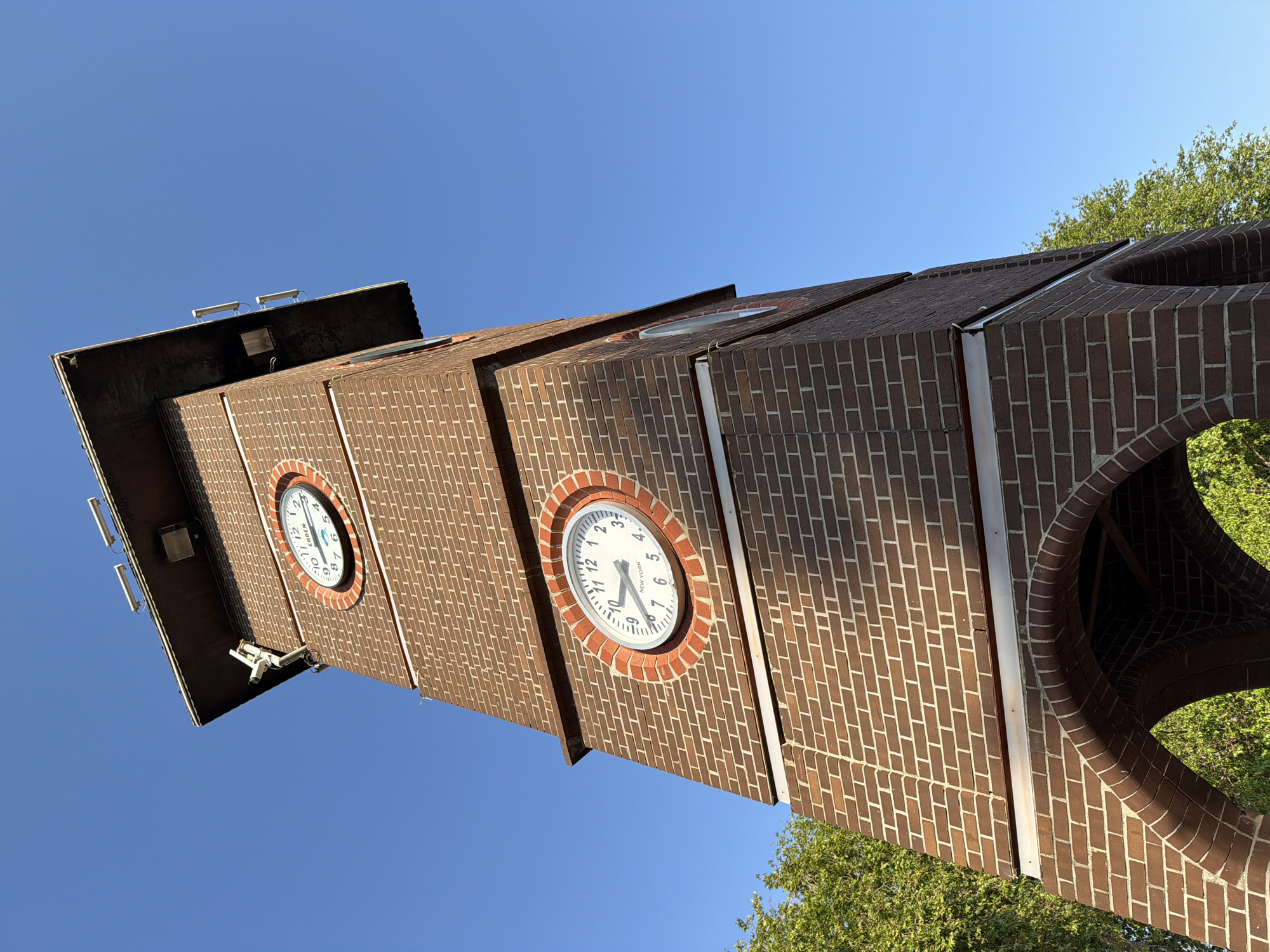 An upward-angled view captures a tall, vertical clock tower constructed from reddish-brown bricks, standing prominently against a clear, bright blue sky. The tower features at least three visible clock faces, each set within a circular, brick-framed opening.
One clock face, centrally located in the image, clearly displays the time as approximately 7:35. Below the '6' on this clock, the text 'NEW YORK' is visible. Another clock face, positioned higher and to the left, shows approximately 10:45 and features the word 'ERBEN'. Both clocks have white faces with black numbers and hands.
The top section of the tower, visible in the upper left of the frame, includes mounted lights and multiple security cameras. An additional security camera is also visible below the 'ERBEN' clock face. Towards the bottom right, an ornate, curved brick archway is part of the tower's structure. The bright sunlight suggests it is daytime with clear weather. Lush green foliage from trees is visible on the right side, framing the tower. Despite one clock indicating 'NEW YORK' time, the tower is located in Alanya, Turkey, suggesting it displays multiple time zones. The perspective emphasizes the tower's height and robust brick construction.