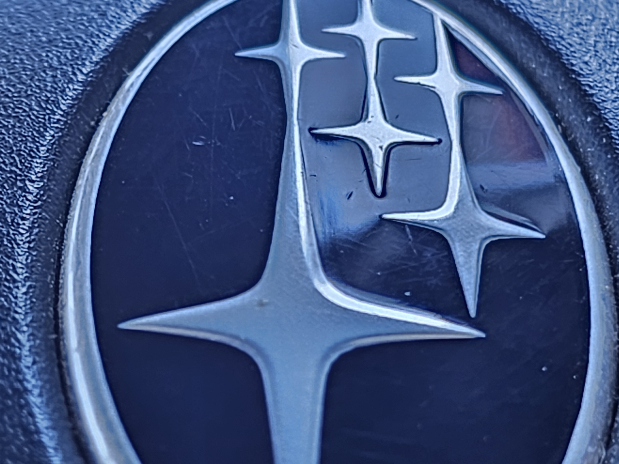 The image is a close-up, macro photograph of the Subaru car emblem. The emblem is silver and set against a dark blue background, possibly the grille or a panel of a car. The emblem features the distinctive Subaru star constellation design, with multiple points of light or stars. The surface of the emblem shows some minor scratches and reflections, indicating it is a metallic surface. The background texture appears to be slightly rough and dark. There is no indication of the city of Nizhny Novgorod, Russia, in the image itself; this information is external to the visual content provided.
