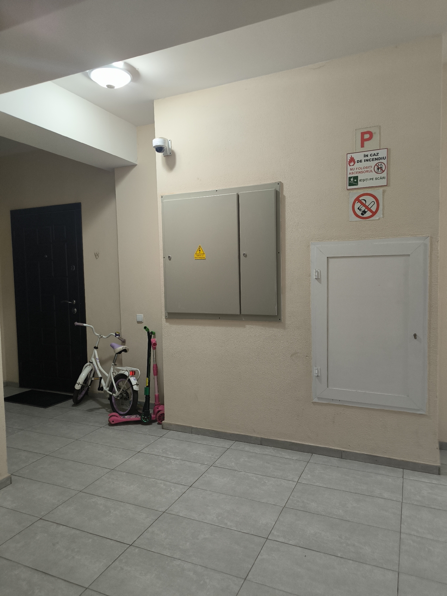 The image depicts an interior hallway in Chișinău, Moldova, likely in a residential building. The walls are painted a light, neutral color, and the floor is covered with large, square gray tiles.
On the left side of the hallway, a black door is visible, with a black doormat in front of it. To the right of the door, a white children's bicycle with purple accents is parked. Next to the bicycle are two scooters: a green and black one and a pink one.
On the wall to the right, a light gray electrical panel with a yellow warning triangle and text in Romanian is mounted. Below that, a white recessed panel with a small lock is installed. Further to the right on the wall, there are two signs. The upper sign is red and white and indicates "P IN CAZ DE INCENDIU NU FOLOSITI ASCENSORUL IEȘIȚI PE SCĂRI," which translates to "P IN CASE OF FIRE DO NOT USE THE ELEVATOR EXIT VIA THE STAIRS." Below this, there is a red and white circular sign with a cigarette crossed out, indicating no smoking.
A security camera is mounted on the wall above the electrical panel. An overhead light fixture illuminates the space. The overall scene suggests a common area in an apartment building.