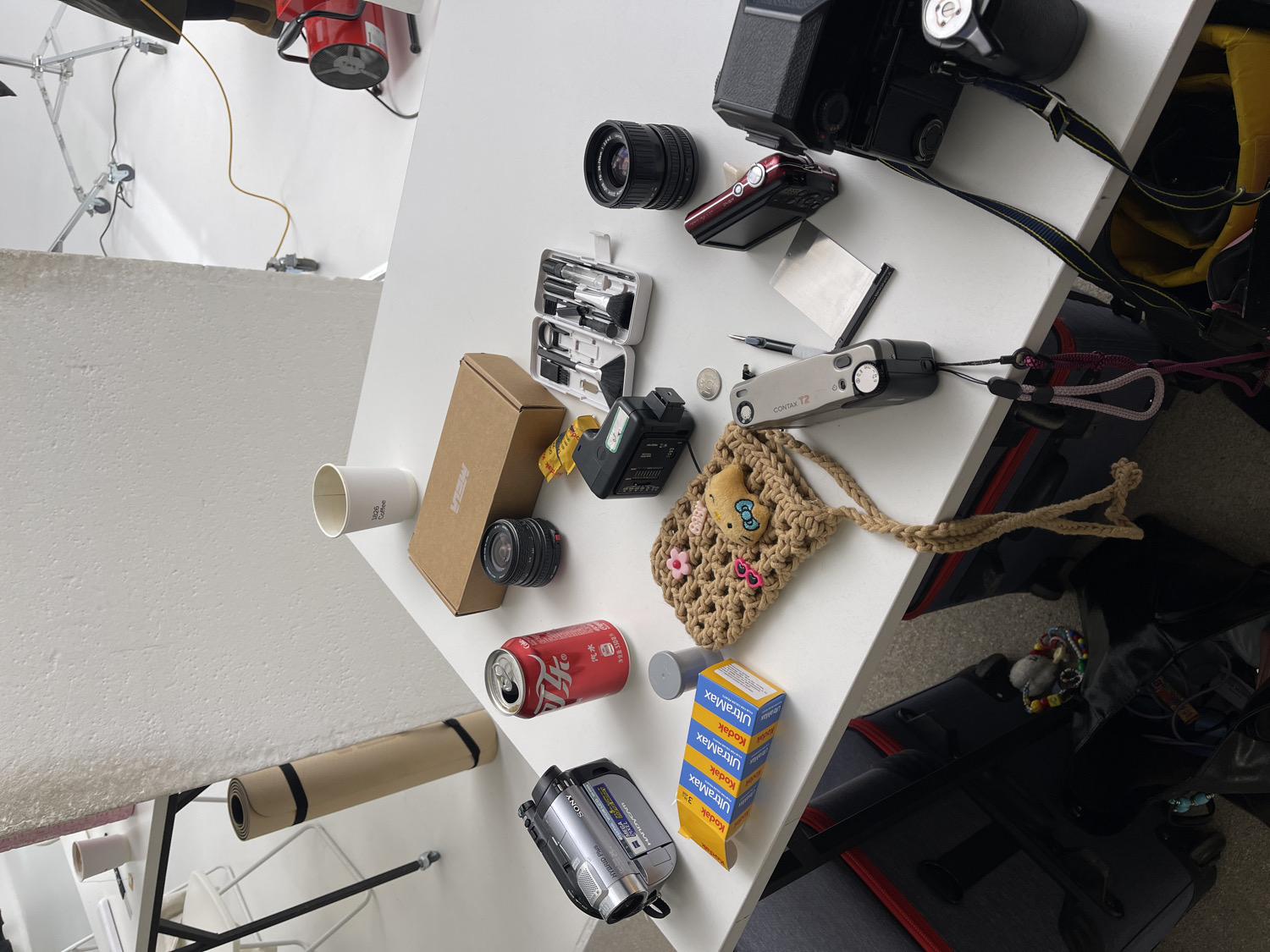 This media file presents a top-down view of a cluttered white tabletop, likely in a photography studio or creative workspace located in Moscow, Russia. The surface is densely packed with a variety of objects, predominantly photography equipment.
Prominent items include several cameras: a large black SLR film camera with a lens, a compact silver "CONTAX T2" film camera with a strap, and a silver "SONY HYBRID Plus" video camera/camcorder. Multiple camera lenses are scattered around, alongside a small black flash unit and a red digital point-and-shoot camera. A white plastic case containing small cleaning tools or brushes is also visible.
Other objects on the table include a stack of three yellow boxes of "Kodak UltraMax" film, a red Coca-Cola soda can with Chinese text "乐 汽水" (meaning "Cola Soda" or "Happy Soda") and "净含量:330" (Net content: 330ml), and a brown cardboard box labeled "MELA". A white paper cup with "bob coffee" printed on its side, a small crocheted pouch featuring a "Hello Kitty" patch, a coin, a pen, a yellow measuring tape, and a reflective silver card complete the arrangement.
The background reveals typical studio elements, such as white walls, a softbox, and a red fan or light fixture. Below the table, a rolled-up light brown mat and a large black bag or case are visible. The scene portrays a functional, lived-in creative environment, suggesting a pause or preparation for a photography session.