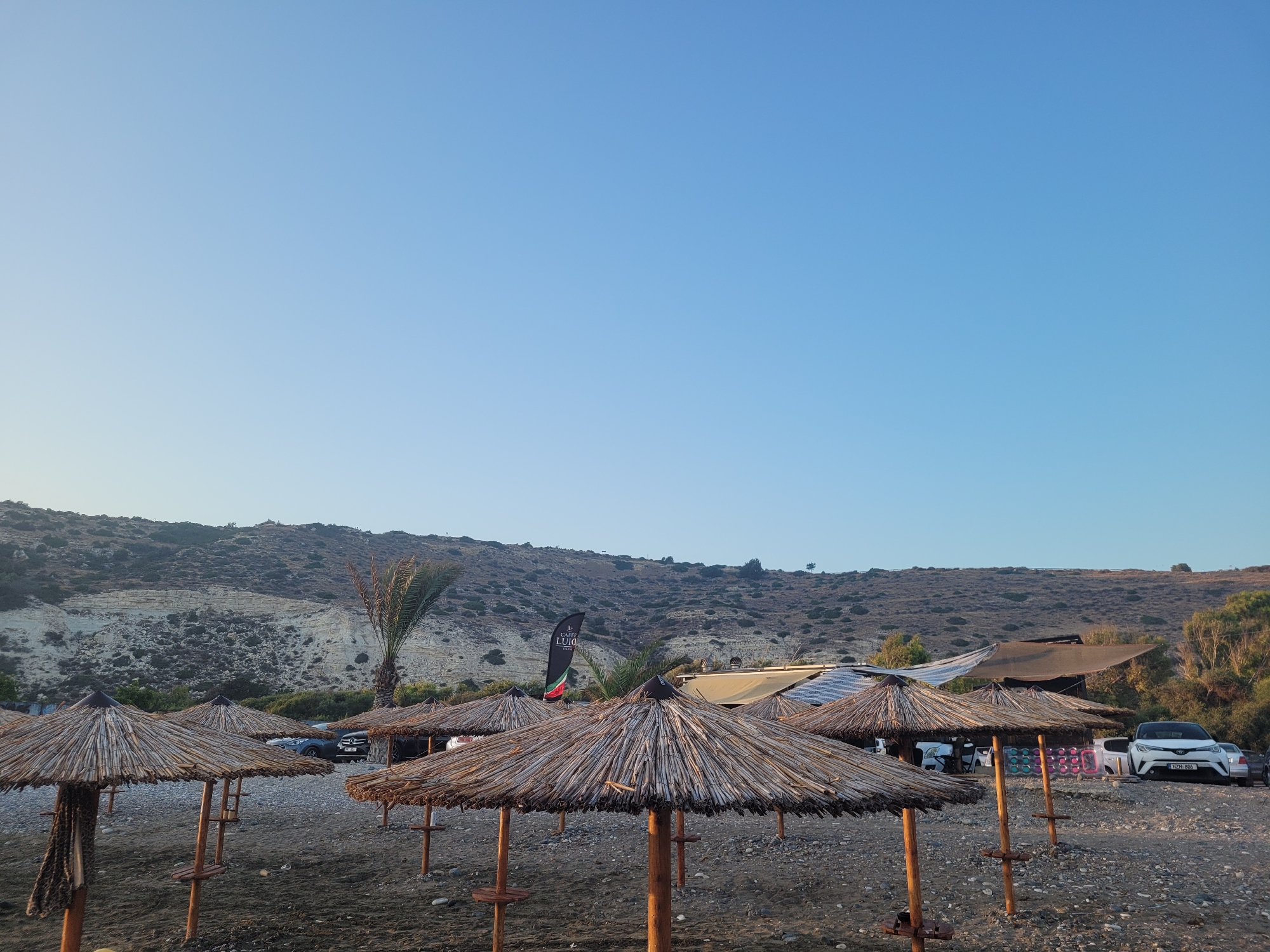 The image depicts a beach scene in Episkopi Lemesou, Cyprus, not the United Kingdom. The foreground is dominated by several thatched umbrellas arranged on a pebble and sand beach. In the mid-ground, there are parked cars, a palm tree, and structures that appear to be part of a beach establishment, possibly a cafe or restaurant, indicated by a flag with the text "CAFFI LUIGI". Beyond the beach, a dry, hilly landscape with sparse vegetation stretches towards the horizon under a clear blue sky. The lighting suggests it is late afternoon or early morning.