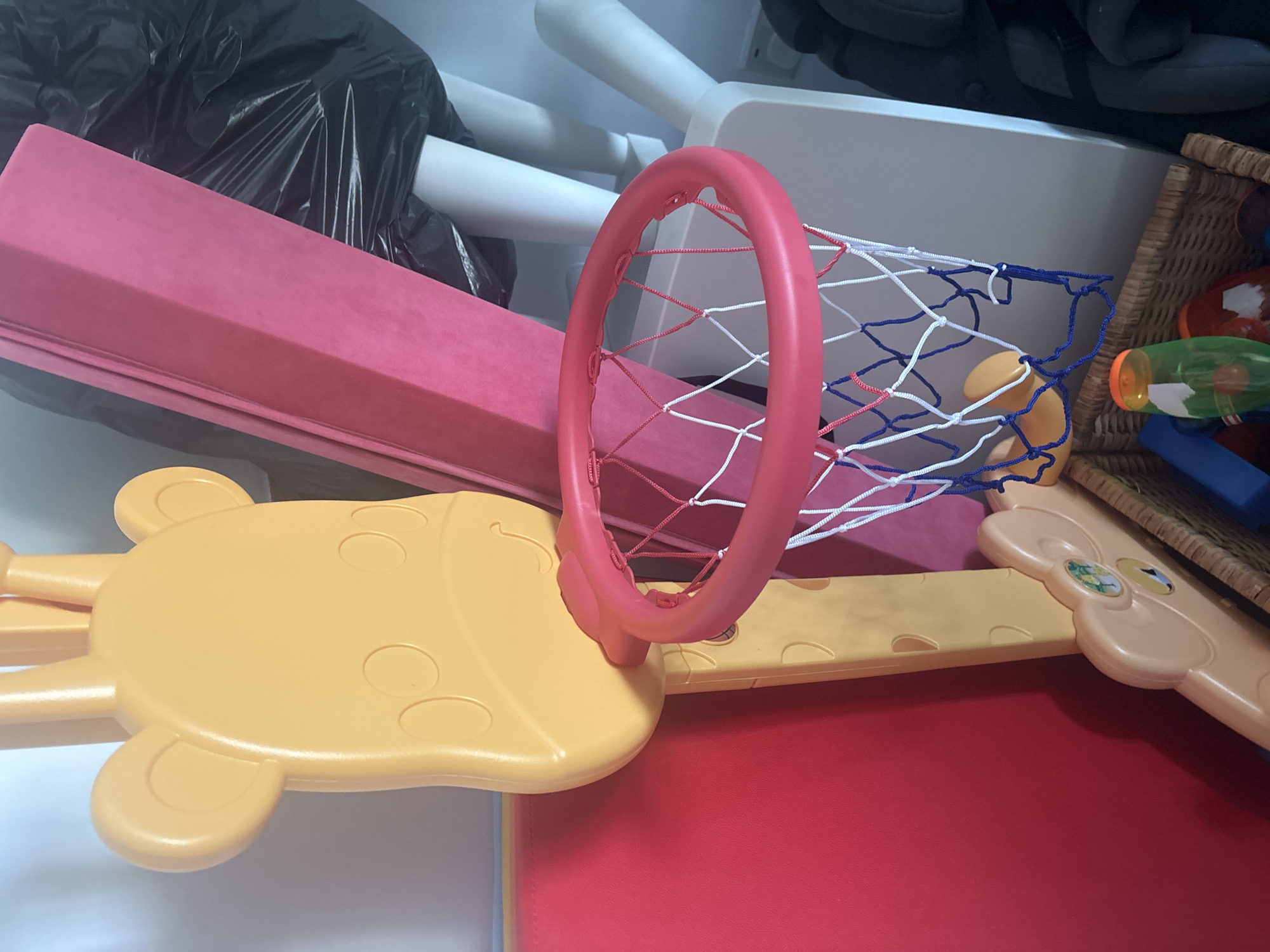 The image shows a close-up of a child's toy basketball hoop, designed to resemble a giraffe's head. The hoop itself is bright pink with a white and blue net. The giraffe's head, made of yellow plastic, has circular brown spots for eyes and oval-shaped ears. Behind the toy, a large pink foam mat is visible, leaning against a black plastic bag and white cylindrical objects. To the left of the basketball hoop, a red mat is partially visible. In the foreground, a wicker basket contains various children's toys, including what appears to be a plastic bottle with an orange lid and a red and white ball. The lighting suggests an indoor setting, possibly a playroom or storage area. The scene is devoid of any context that would place it specifically in Dubai, United Arab Emirates.