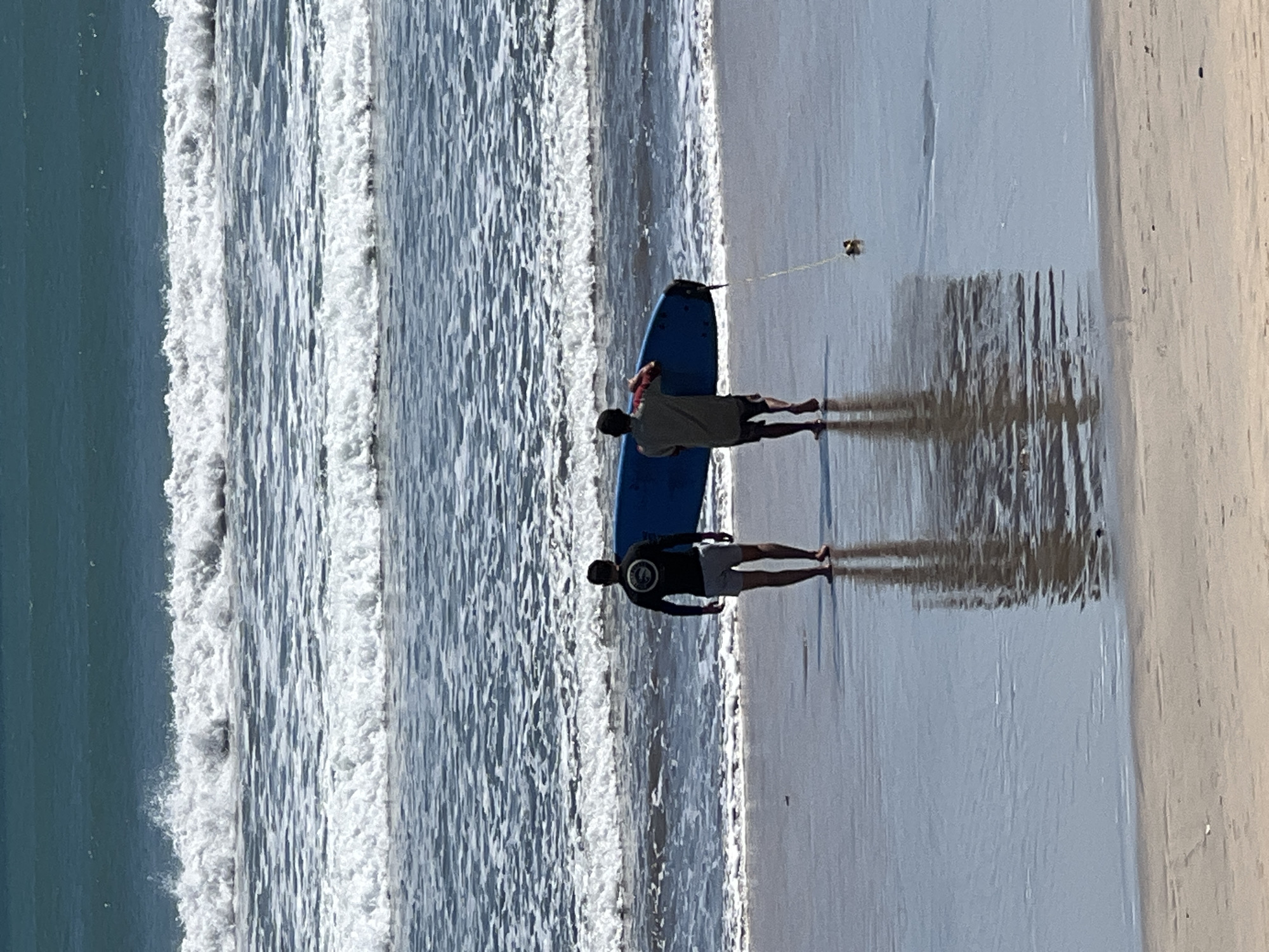 Two individuals are walking on a wet sandy beach towards the ocean in Kuta, Indonesia. The person on the left, wearing a black long-sleeved shirt and light-colored shorts, is facing away from the camera. The person on the right, also facing away, is carrying a blue surfboard under their arm and wears a short-sleeved shirt and dark shorts. A thin yellow leash is attached to the surfboard and trails behind it on the wet sand. The ocean in the background displays several lines of white-capped waves under a bright sky. The wet sand reflects the figures and the sky.
