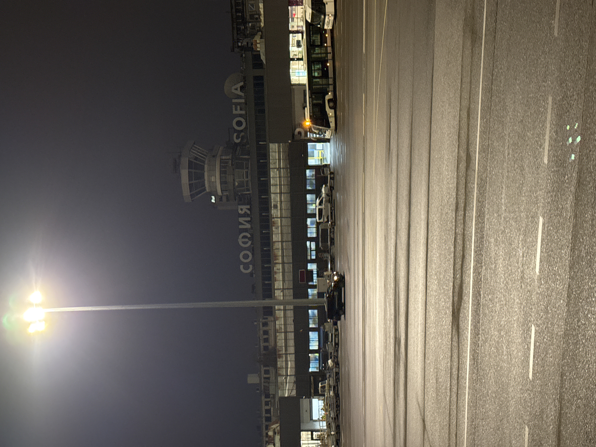 The image shows Sofia Airport at night. The airport terminal building is visible with "SOFIA" written vertically on its facade. An air traffic control tower stands nearby. A row of vehicles, including buses, is parked in front of the building. The foreground features a paved area with marked lines, illuminated by a tall light pole on the left. The sky is dark, indicating nighttime.