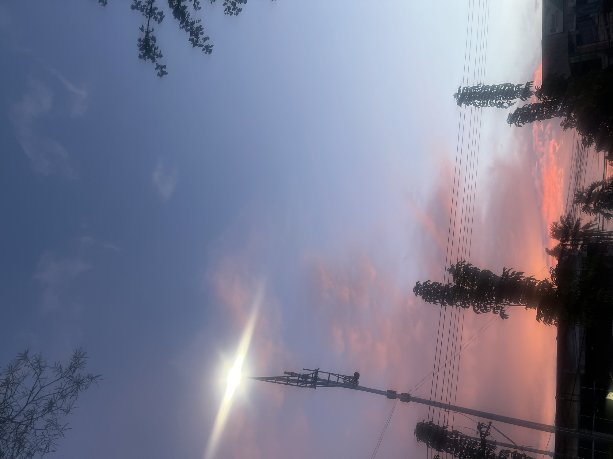 A serene twilight sky dominates the frame, transitioning from a soft lavender and pink hue at the horizon to a deep blue overhead. Wispy clouds are scattered throughout, tinged with the warm colors of sunset. In the foreground, silhouetted against the vibrant sky, are several tall, slender trees with dense foliage, their shapes suggesting they are climbing structures or vines.
To the left, a utility pole rises, its structure visible, topped by a bright, luminous lamp that beams a strong white light diagonally across the sky, creating a lens flare effect. Several horizontal power lines stretch across the middle of the image, connecting to other unseen poles.
At the very bottom of the frame, the dark silhouettes of buildings and more trees can be discerned. The overall impression is one of a peaceful evening scene in an urban or semi-urban environment. Given the context of Banda Aceh, Indonesia, this scene likely depicts a street or open area in the city during dusk, where modern infrastructure like utility poles and buildings coexist with lush tropical vegetation under a beautiful sky.
