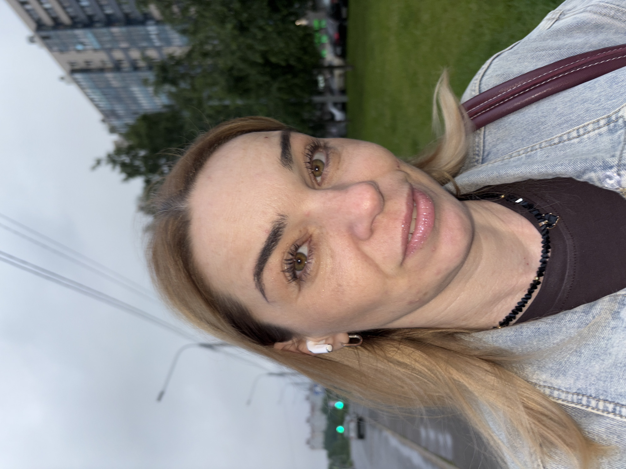 A medium shot shows a woman with blonde hair and green eyes, wearing white earbuds and a denim jacket, looking into the camera. She has on a dark necklace with black beads and a gold pendant. The woman is outdoors in what appears to be a city. In the background, a multistory building and trees are visible. A street with cars and streetlights can be seen in the distance. The sky is overcast. The location is identified as Unknown, Russia.