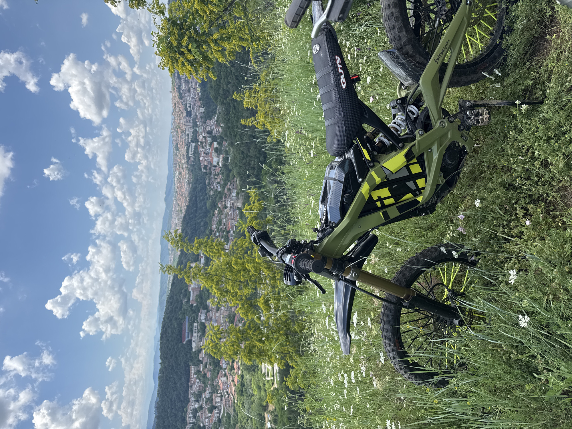 An electric dirt bike, predominantly green with yellow accents, is parked on a grassy, flower-filled hillside overlooking the city of Veliko Tarnovo, Bulgaria. The bike is positioned in the foreground, with its front wheel slightly turned. In the background, the cityscape of Veliko Tarnovo unfolds, characterized by its distinctive red-tiled roofs and a hilly terrain. The sky above is blue with scattered white clouds. The environment is natural and semi-urban, with lush vegetation and a panoramic view of the city and surrounding hills.