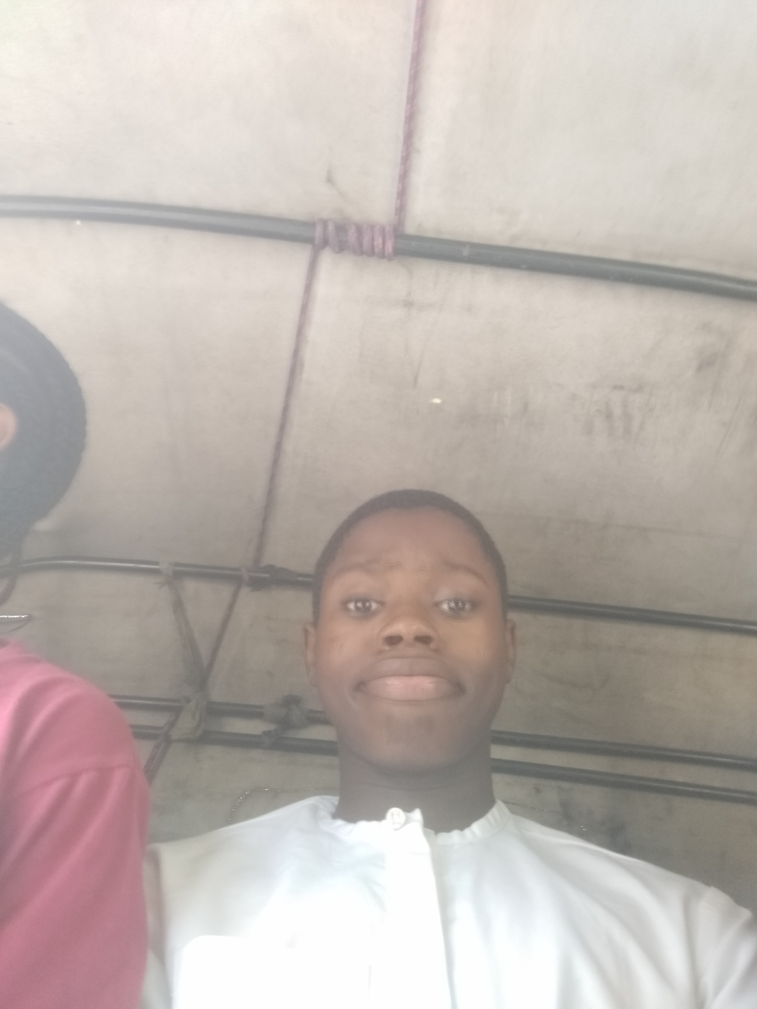 A medium shot depicts a young Black male wearing a white, collared shirt, seemingly inside a vehicle. To the left, a portion of another individual is visible wearing a red shirt and a black hat. The ceiling of the vehicle is visible, showing metal bars and a rope tied together. The image was taken in an unknown location within the city of Unknown, Unknown.