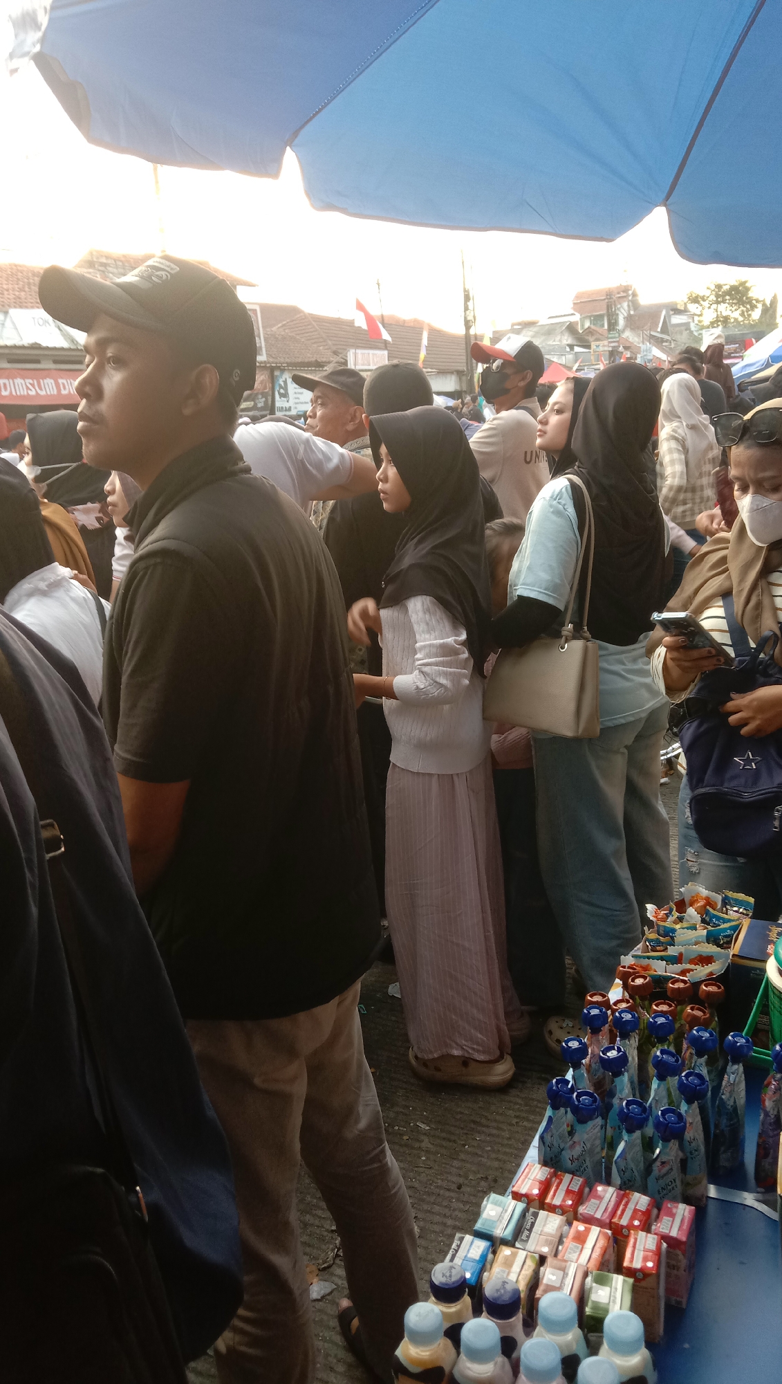In Pesantren, Indonesia, a street market scene unfolds under a large blue canopy. A man in a black t-shirt and baseball cap stands prominently in the foreground, his profile visible as he looks to the left. Behind him, a crowd of people are visible, many wearing hijabs. One woman in a light pink dress and hijab stands out, her gaze directed forward. To her right, another woman in a light blue shirt and jeans, also wearing a hijab, holds a light beige handbag. A few people are wearing masks. In the lower right corner, a table is laden with various packaged food items, including small boxes of cigarettes and bottles of drinks, suggesting a vibrant commercial activity. The lighting suggests it is daytime, possibly late afternoon, with sunlight filtering through the canopy. The ground appears to be paved with asphalt. The overall atmosphere is one of a bustling market, with numerous individuals engaged in shopping or socializing.