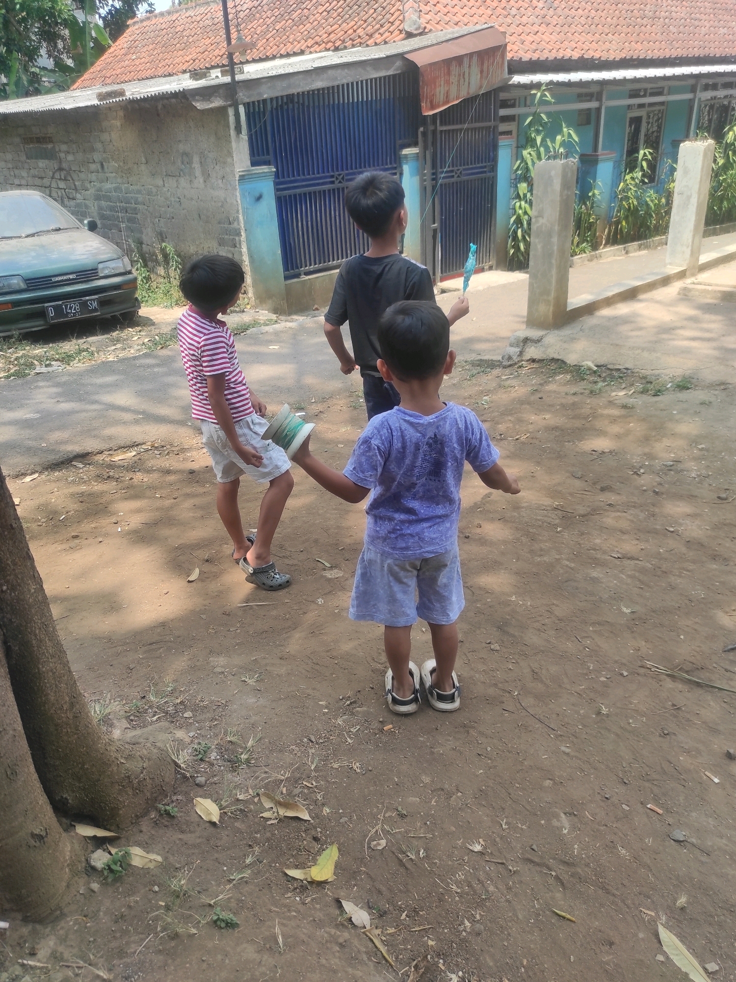 Three children are engaged in play in front of a blue and grey residential building in Padalarang, Indonesia. The child in the foreground, facing away from the camera, wears a purple t-shirt and light blue shorts. To the left, a second child in a striped pink and white shirt and white shorts stands with their back to the camera, holding a cylindrical object with string wrapped around it. A third child, also facing away, stands in the middle ground wearing a black t-shirt and holding a blue toy. A green car is parked to the left of the building, and the ground is a mixture of dirt and sparse vegetation. The building has a red tiled roof and a blue metal gate.