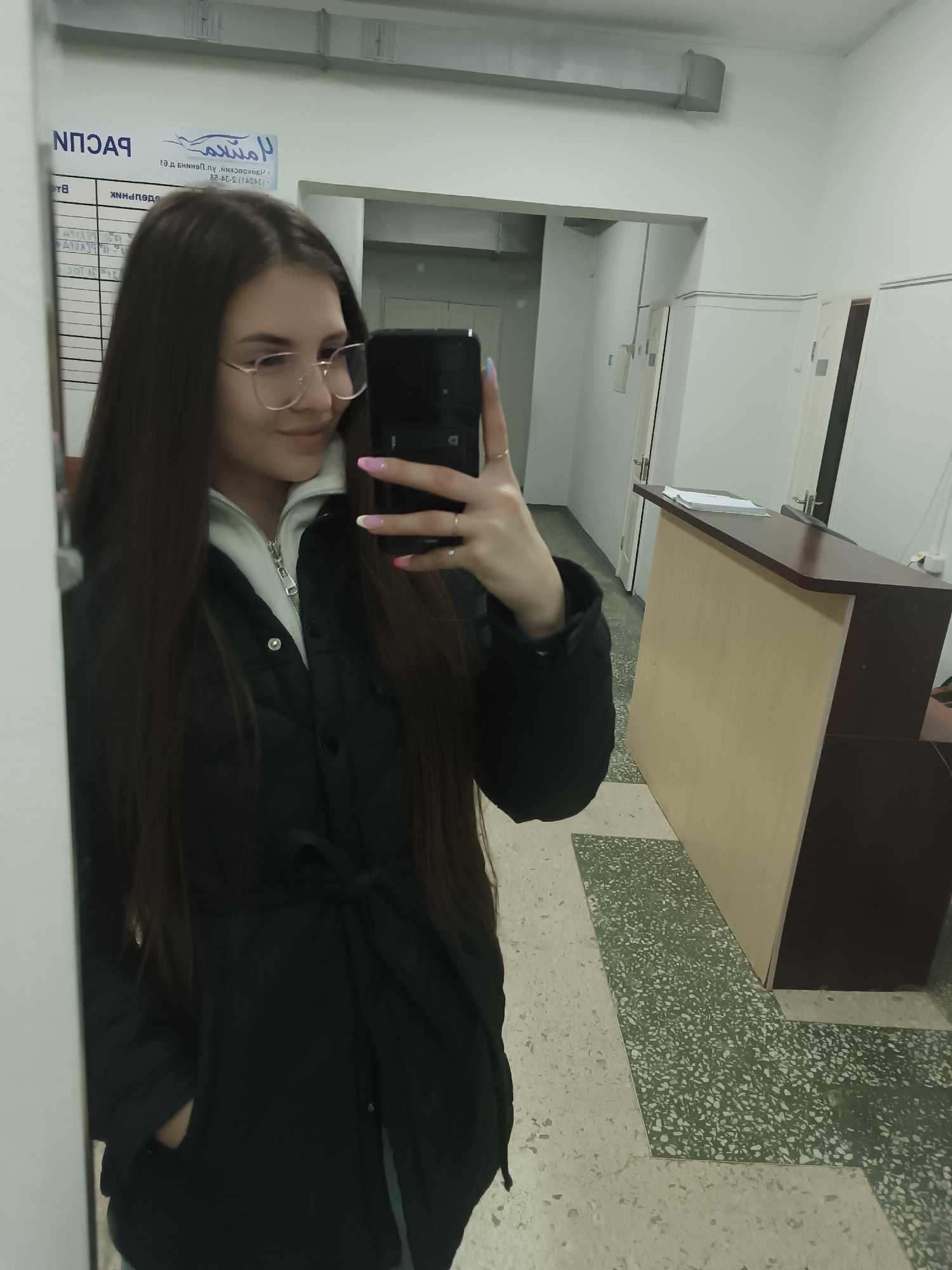 A person stands in front of a mirror, taking a selfie with a smartphone. The individual is a young woman with long, straight, dark brown hair and wears glasses with gold frames. She has on a black, quilted jacket with a tie belt and a white zip-up hoodie underneath. Her fingernails are painted with different pastel colors.
The background shows a reception area. A reception desk with a light wood-colored front and a dark brown top is visible, and there's paperwork on top. In the background, there are white doors and walls. The floor has a speckled green and white pattern. There is a sign hanging on the wall to the left, with Russian text. The setting appears to be an office or medical facility. The location is the city of Unknown, Unknown.