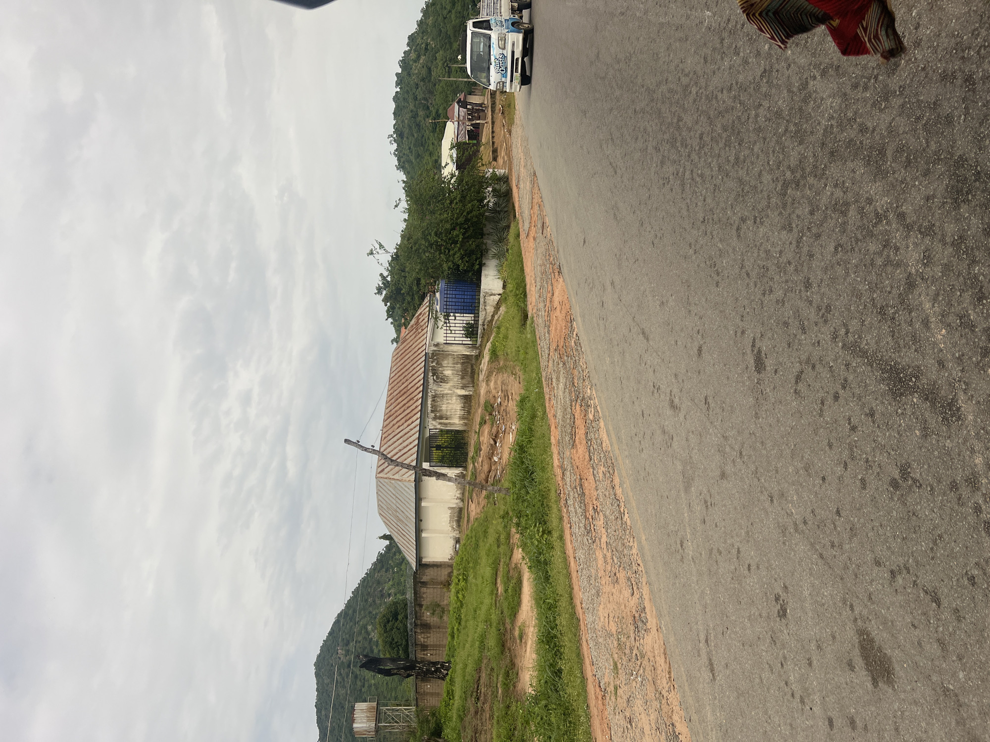 A horizontal shot shows a road beside a building and a hill. The road is paved with asphalt and has some loose dirt on the side. The building has a corrugated metal roof and a light-colored facade. There is a blue water tank in front of the building. Behind the building, a lush green hill rises, covered in trees. The sky is overcast, suggesting a cloudy day. In the foreground, a person's legs and colorful clothing are visible, suggesting they are walking on the road. Further down the road, a white truck with blue markings is driving. The scene appears to be in a rural or semi-rural area, possibly in Nigeria, given the context. The overall mood is mundane and everyday. No text is clearly visible in the image.