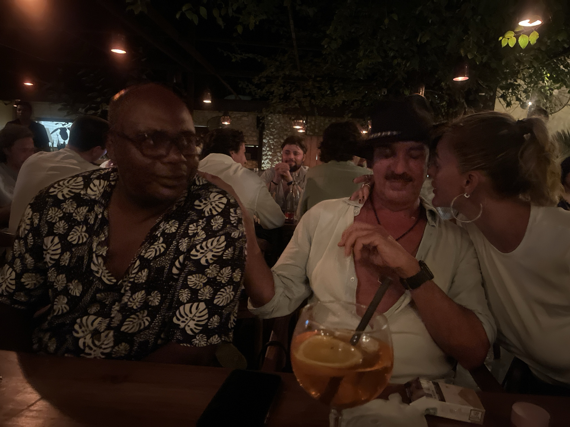 The image was taken at night in what appears to be a restaurant in Kuta Selatan, Indonesia. The lighting is dim, and several people are seated at tables.
In the foreground, there are three people. On the left is a Black man wearing a dark, short-sleeved shirt with white floral patterns and glasses. He has a shaved head. To his right is a light-skinned man wearing a cowboy hat and a light-colored button-down shirt. He has a mustache, a necklace, and a watch. He is sitting next to a woman wearing a white shirt. The woman is whispering into the man's ear and has her hair in a ponytail. The man has a drink in a glass with a straw and an orange slice. A package of cigarettes is on the table near the glass.
In the background, several other people are seated at tables, some facing the camera and some with their backs to it. Overhead, there appear to be trees with lights hanging down, creating a dimly lit and intimate atmosphere. The rest of the restaurant is visible in the background with other patrons and staff visible. A cell phone is on the table.
