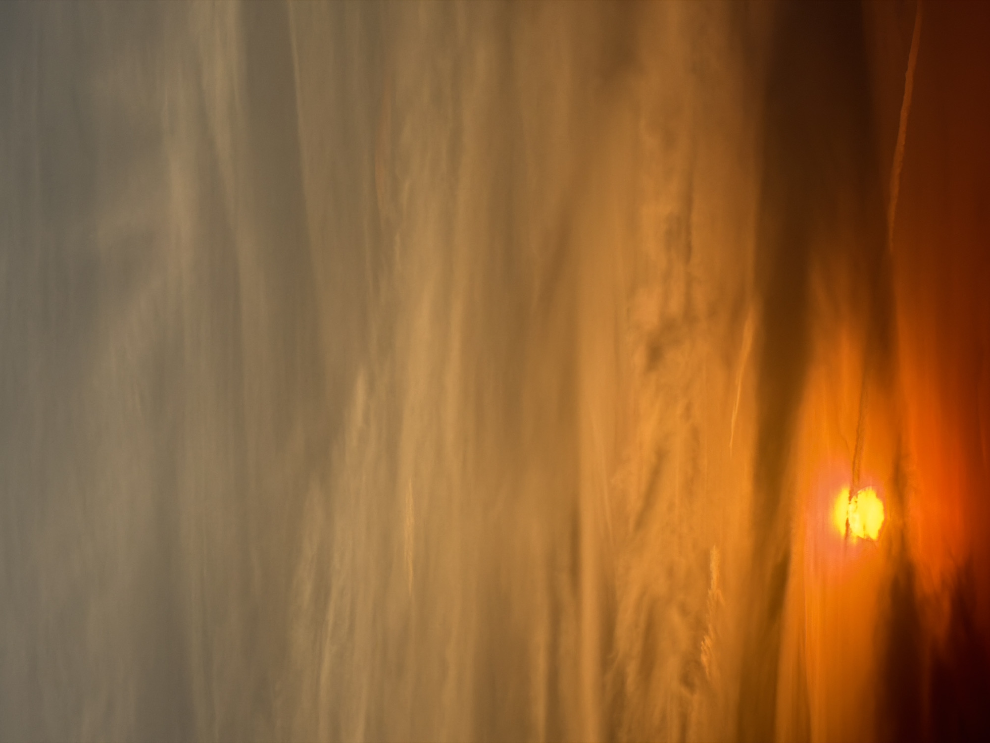 The image captures a sunset in Unknown, Unknown, featuring wispy clouds that transition from grey to orange hues. The sun, a bright orange orb, is partially obscured by the clouds on the right side of the frame. A contrail is visible towards the upper-right corner.