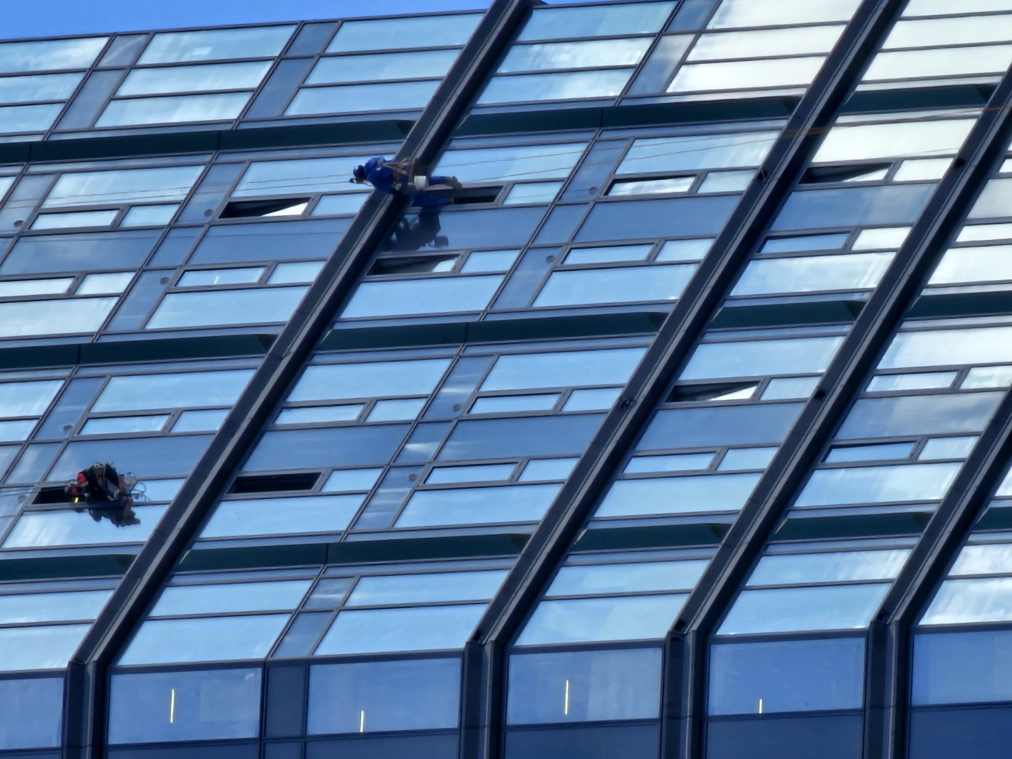 The image depicts two individuals engaged in window washing on the exterior of a modern, glass-facaded building. One person, dressed in a blue uniform and suspended by ropes, is positioned higher on the building's surface. They appear to be actively cleaning the windows. Below this individual, another person, wearing a red and black outfit, is also suspended and working on the windows. The building's facade is composed of numerous rectangular glass panels, some of which are open or angled, reflecting the bright blue sky and scattered clouds. The scene suggests a daytime activity under clear weather conditions. The architecture of the building, with its angular design and extensive use of glass, is a prominent feature. While the specific location is provided as Moscow, Russia, no explicit text or overt cultural cues are visible within the image itself to confirm this. The activity highlights the challenging and potentially dangerous nature of high-rise window cleaning.