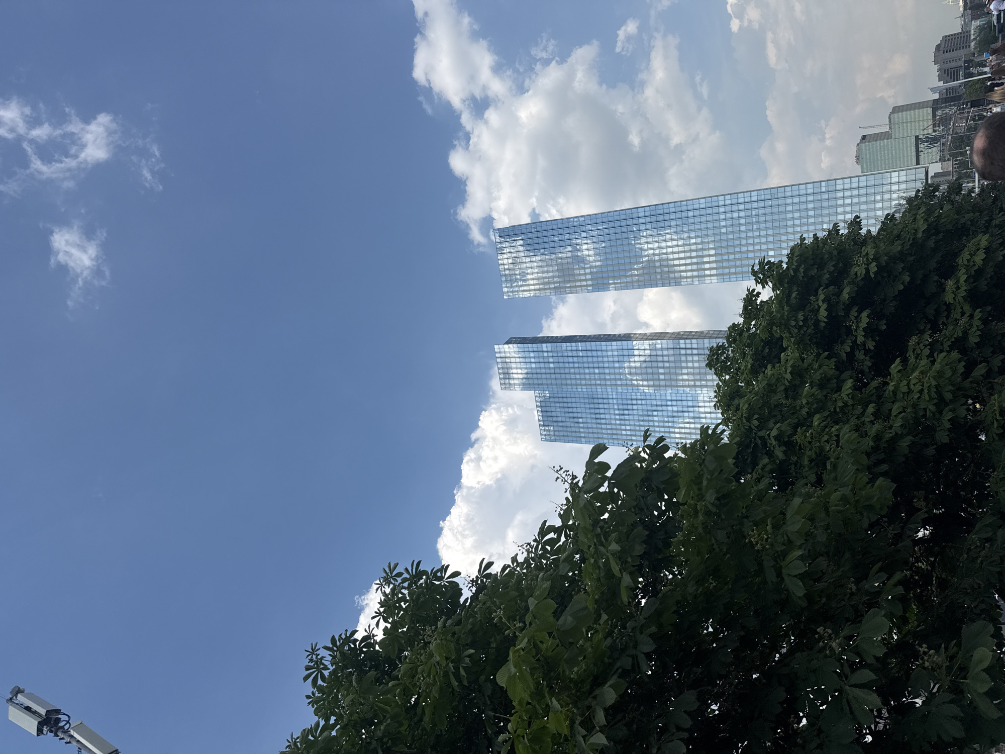 The image shows two modern skyscrapers with glass facades reflecting the sky and clouds. The buildings appear to be made of many rectangular panels, giving them a sleek and contemporary look. In the foreground, lush green trees with full foliage partially obscure the view of the skyscrapers. The sky is a vibrant blue, dotted with fluffy white clouds, suggesting a pleasant day.
To the left of the frame, a portion of what appears to be telecommunications equipment or a cell tower is visible at the bottom. The scene is shot from a low angle, looking upwards towards the sky and the tops of the buildings. The overall impression is one of urban development contrasted with nature. The reflective surfaces of the skyscrapers create an interesting interplay between the man-made structures and the natural elements of the sky and trees. The lighting suggests it is daytime, likely mid-morning or afternoon, given the brightness and the blue sky. No specific event or activity is depicted, rather it is a static view of the city landscape.