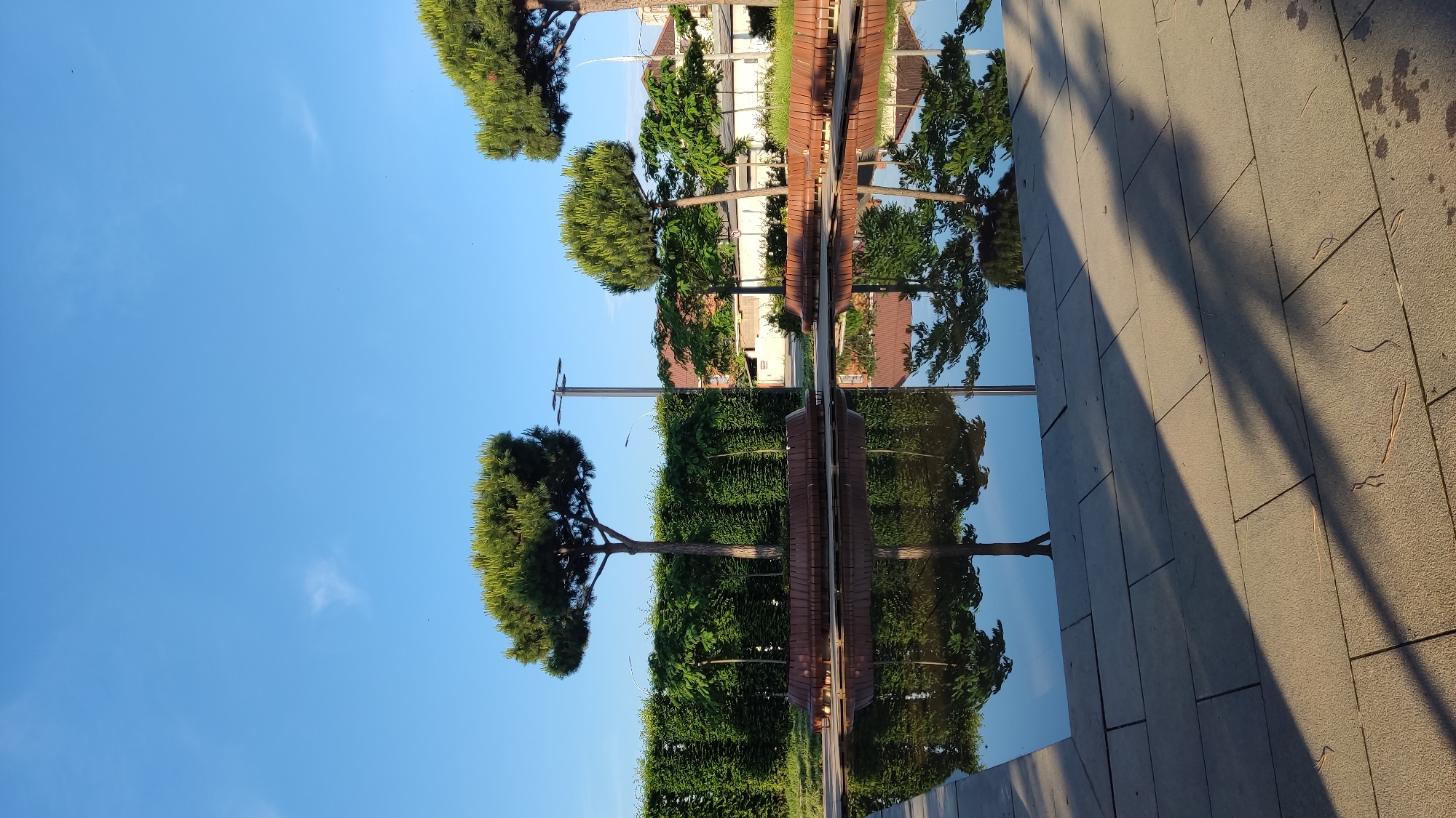 The image depicts a tranquil park scene under a clear blue sky in Krasnodar, Russia. In the foreground, a rectangular body of water reflects the surrounding elements. The water's surface is smooth, acting as a mirror to the trees and the sky.
Beyond the water, a neatly trimmed hedge forms a verdant backdrop. Several umbrella-shaped pine trees stand tall in front of the hedge, their green canopies reaching towards the sky. Between the trees, wooden benches are arranged, appearing to be part of a continuous seating structure. A lamppost is visible behind the trees, its dark pole extending upwards.
In the mid-ground, the reflections of the trees, benches, and the sky are clearly visible in the water. The water itself is contained within a paved area made of large, rectangular stones.
Further in the background, more trees and buildings can be seen, hinting at the urban setting. The buildings have tiled roofs, typical of residential or commercial structures. The overall impression is one of a well-maintained public space, likely a park or a promenade, designed for relaxation and enjoyment of nature within the city. The lighting suggests it is daytime, possibly morning or late afternoon, given the long shadows cast on the paved area. No people are present in the image.