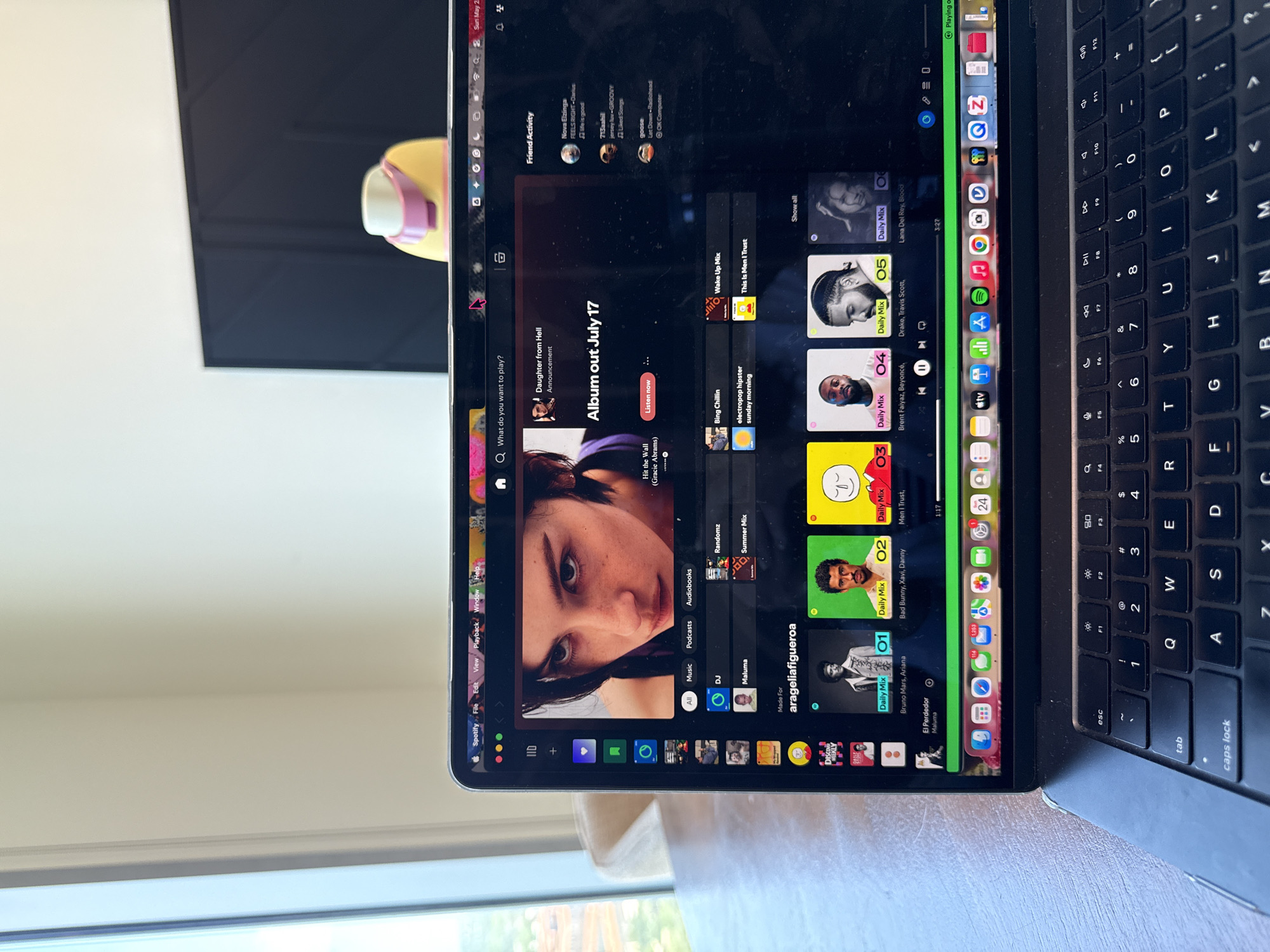 The image shows a MacBook laptop displaying the Spotify application. The screen is filled with the Spotify interface, including album art, playlists, search bar, friend activity, and the dock. The laptop's keyboard is visible. The background includes a glimpse of the room, showing sunlight, a window view, and a desk. The laptop is positioned indoors. The image does not contain information to determine it is in Austin, Texas, USA.