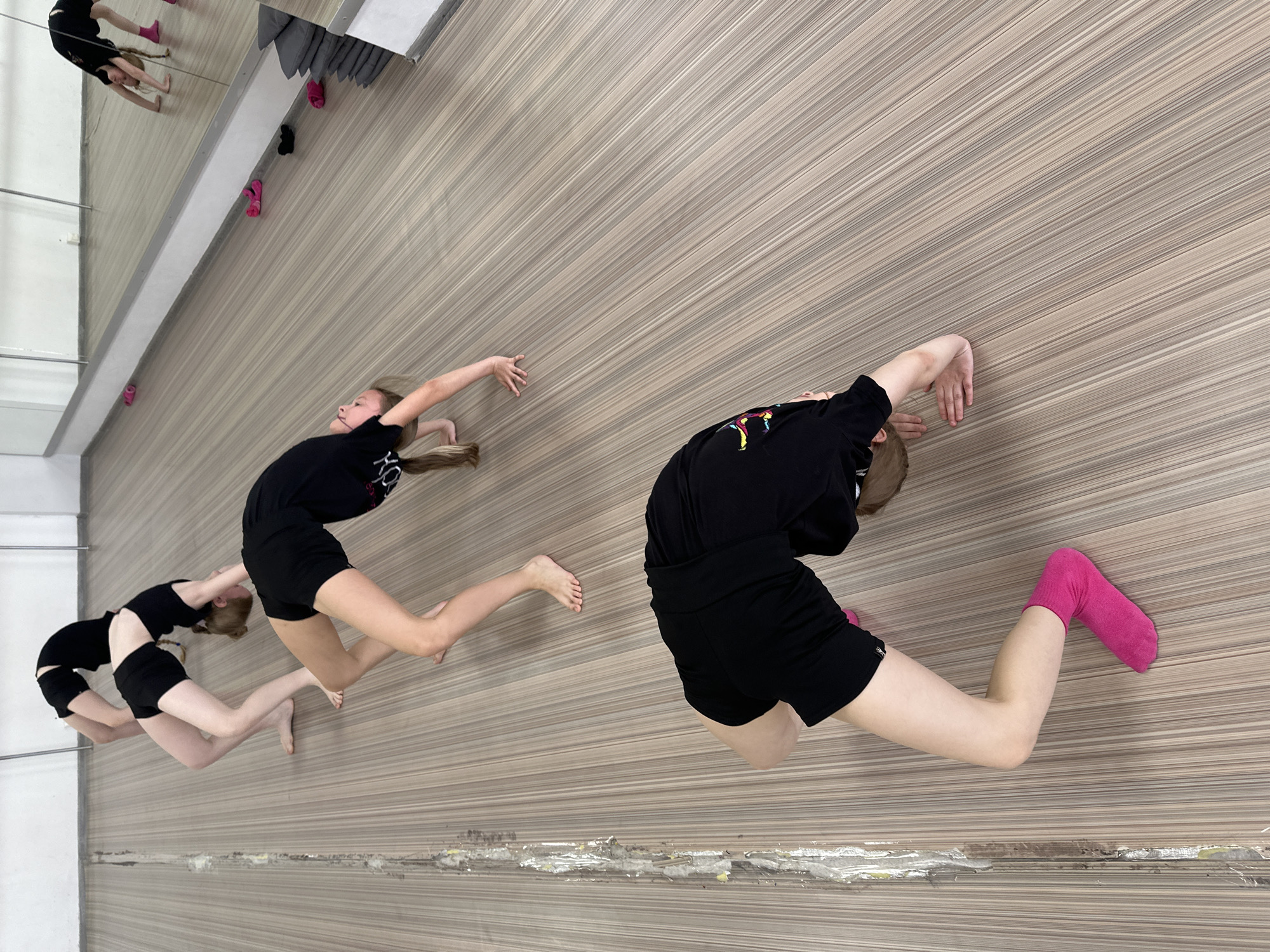 Three girls, possibly gymnasts or dancers, are performing arched poses in a mirrored studio. The mirrored wall is on the left, and the other wall has vertical wood paneling. All are wearing dark-colored athletic clothing and are barefoot, with one wearing pink socks. The floor appears to be light-colored and potentially damaged along the bottom edge. There are pink and black socks near the mirror. It is presumed that the image was taken in Votkinsk, Russia, but there are no explicit indicators in the image.
