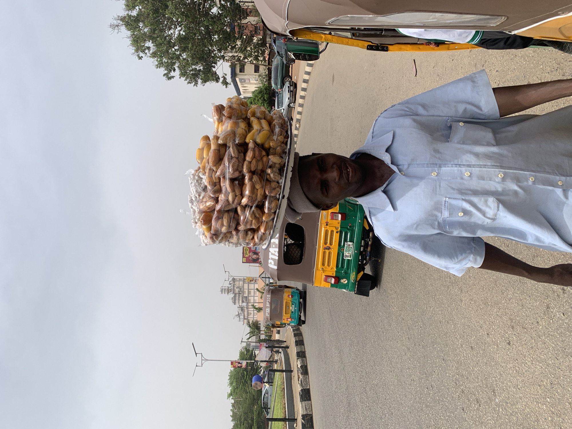 This image captures a vibrant street scene, likely in Jos, Nigeria, showing a male street vendor carrying a large tray of goods on his head. The man, with dark skin, wears a light blue short-sleeved collared shirt and a light-colored cap or cloth on his head. He has a slight smile on his face, looking towards the viewer.
The sizable, round, silver-colored tray balanced skillfully on his head is filled with numerous individually packaged food items. These appear to be a variety of dried fruits, possibly dates, and small baked goods or snacks, mostly wrapped in clear plastic.
The setting is an urban or semi-urban street. A paved road stretches into the background, flanked by a black and white striped curb or median separating it from a grassy area with trees. Several vehicles are present on the road: part of a light brown car or auto-rickshaw is visible in the foreground, and behind the vendor, a green and yellow auto-rickshaw (keke Napep) is clearly seen from the rear. Its license plate displays "BA-909K" and likely "PLATEAU" below it. Another brown auto-rickshaw with "PTA-1095" on its side is further down the road, and other cars are parked on the left. Buildings can be seen in the distance, and a large tree with green foliage is prominent in the upper left.
The activity depicted is street hawking, a common practice where vendors sell goods directly on the streets. The time of day is bright daylight, indicated by the clear, light sky and strong illumination, suggesting sunny weather. A yellow billboard with "Yes!" is visible in the far background.
