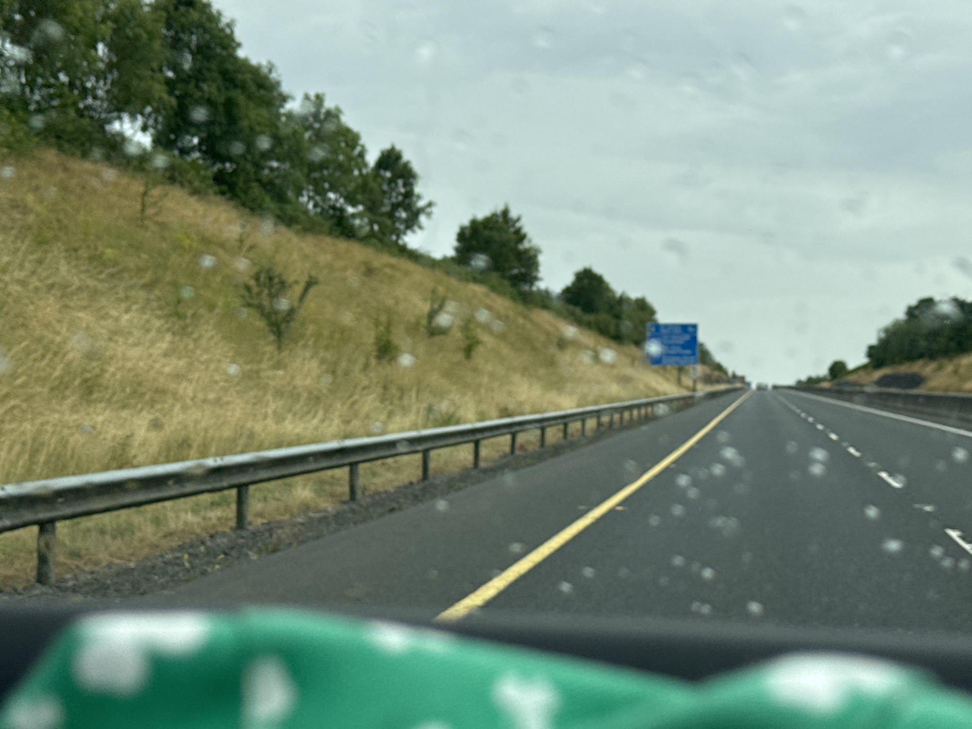 The image depicts a view from inside a vehicle traveling on a highway. The highway has a yellow line dividing the lanes and white dashed lines indicating lane separation. A metal guardrail runs along the left side of the road, separating it from a grassy embankment covered in dry, yellow grass and scattered trees. The sky is overcast and appears to be raining, with water droplets visible on the windshield. In the distance, a blue highway sign is visible on the right side of the road. The foreground shows a blurred green object, possibly a piece of fabric or a dashboard element. The scene appears to be a typical highway setting, likely in Ireland given the mention of Bennettsbridge, Ireland.