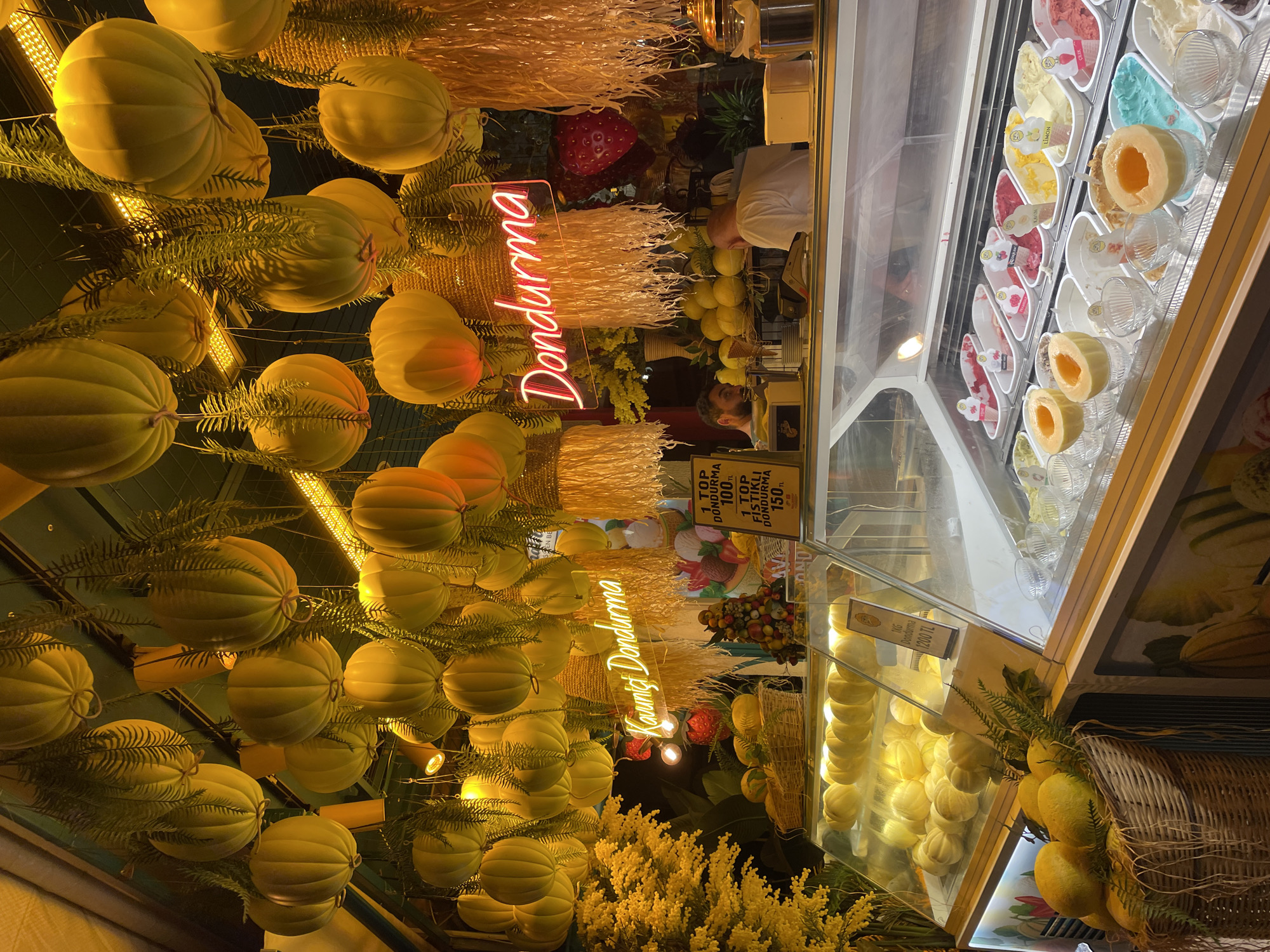 This is a medium shot of an ice cream shop in Kadıköy, Turkey, at night. The shop is decorated with numerous yellow, pumpkin-shaped ornaments hanging from the ceiling, interspersed with artificial fern branches. A neon sign reading "Dondurma" in pink letters hangs prominently. In front of the shop, a display case filled with various flavors of ice cream and sliced cantaloupes is visible. To the left, a basket overflowing with yellow fruits, possibly lemons or small melons, rests near a display of whole cantaloupes. Two men are working behind the counter. One man, partially visible on the left, appears to be interacting with a customer or preparing an order. The second man, on the right, is facing away from the camera and is also behind the counter. The overall atmosphere is warm and inviting, with soft lighting illuminating the colorful ice cream display and the festive decorations.