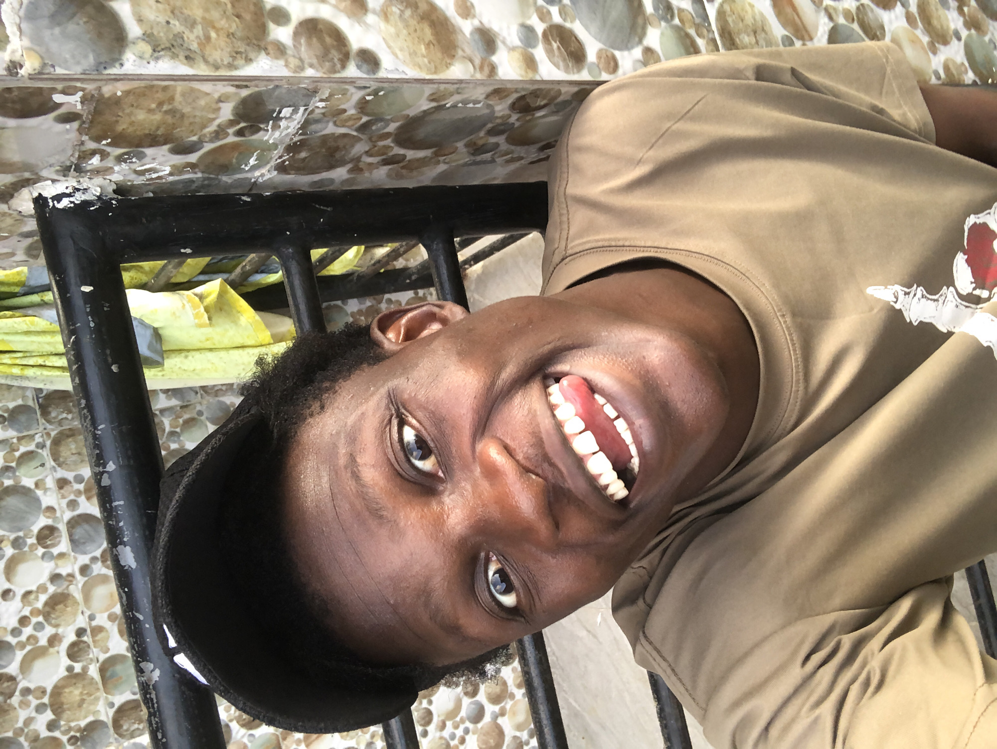 A young Black male wearing a black baseball cap and a tan t-shirt with a white graphic on the chest is captured in a close-up, selfie-style portrait. He has dark, curly hair and is sticking his tongue out while smiling broadly. The background consists of a tiled wall with circular, stone-like patterns and a black metal railing. This appears to be a casual outdoor setting, possibly in Ado, Nigeria.