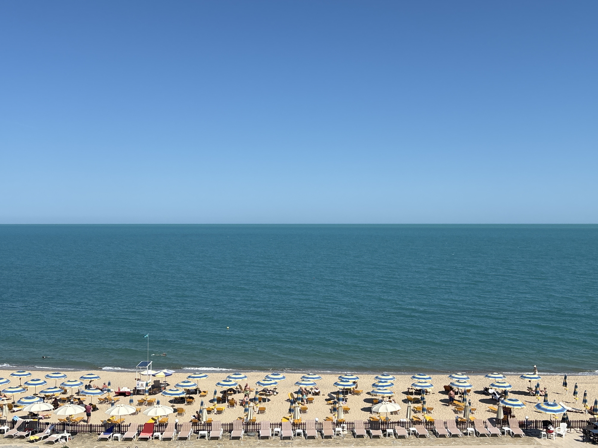 This is a high-angle, unobstructed, eye-level view of a beach in Varna, Bulgaria on a clear, sunny day. The vast, turquoise sea stretches to the horizon, meeting a cloudless, pale blue sky. The sandy beach is lined with rows of lounge chairs and umbrellas, predominantly striped blue and yellow, with some solid white. Several beachgoers are visible, some seated on chairs, others standing or walking on the sand. A lifeguard stand with a green flag is positioned near the water's edge. The scene conveys a sense of leisure and summer vacation.