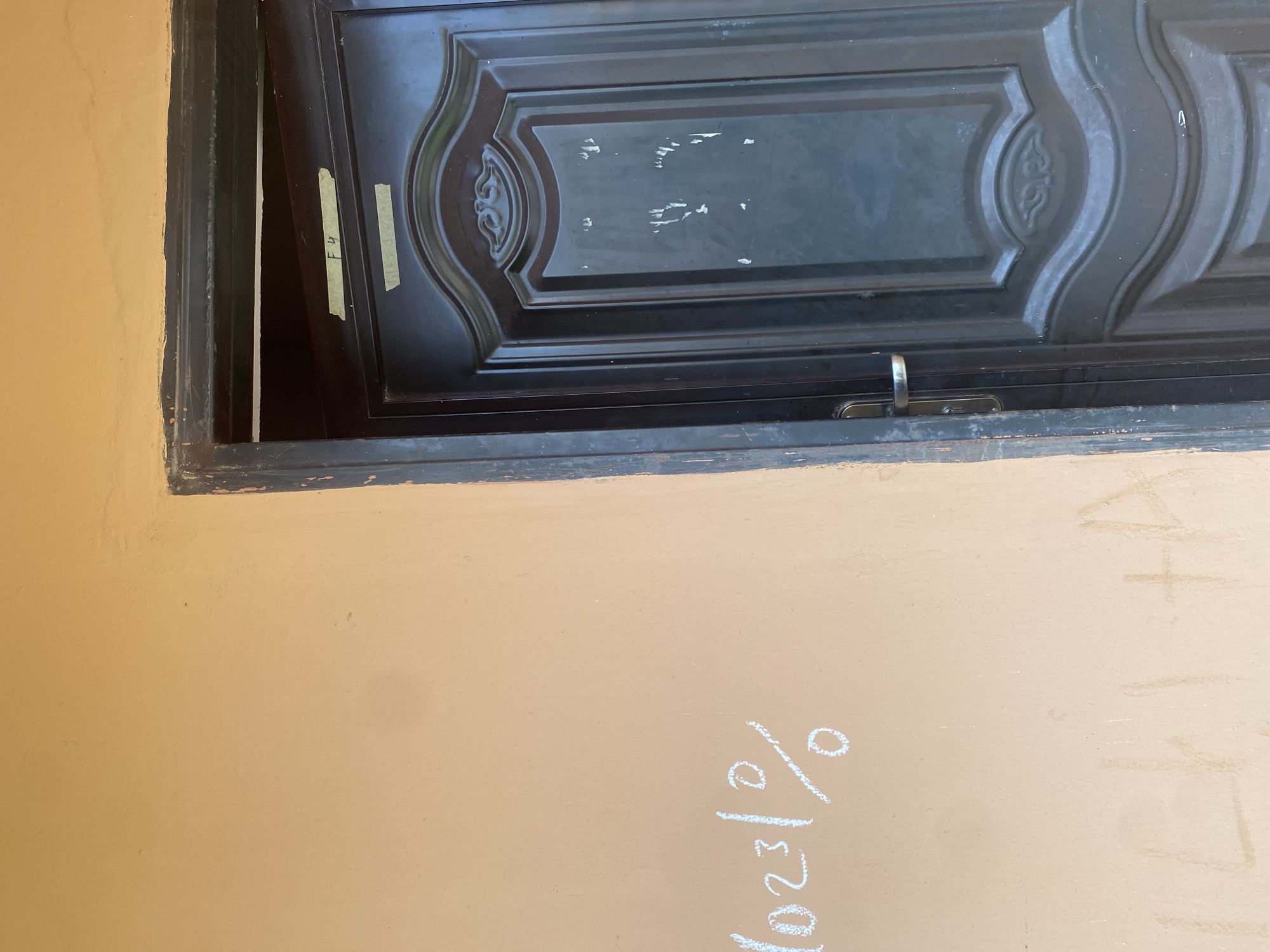The image shows a closed, dark gray metal door on the right side of the frame. The door has an ornate design in the upper portion and a silver handle on the left side. Two pieces of yellow tape with writing are affixed to the door near the top. On the left side of the frame, on an orange wall, is writing in white chalk. The chalk writing appears to be a series of numbers and a percentage sign, "10231%". Faint chalk markings, possibly letters, are also visible lower down on the wall. The environment appears to be an outdoor wall in Bulamare, Nigeria.