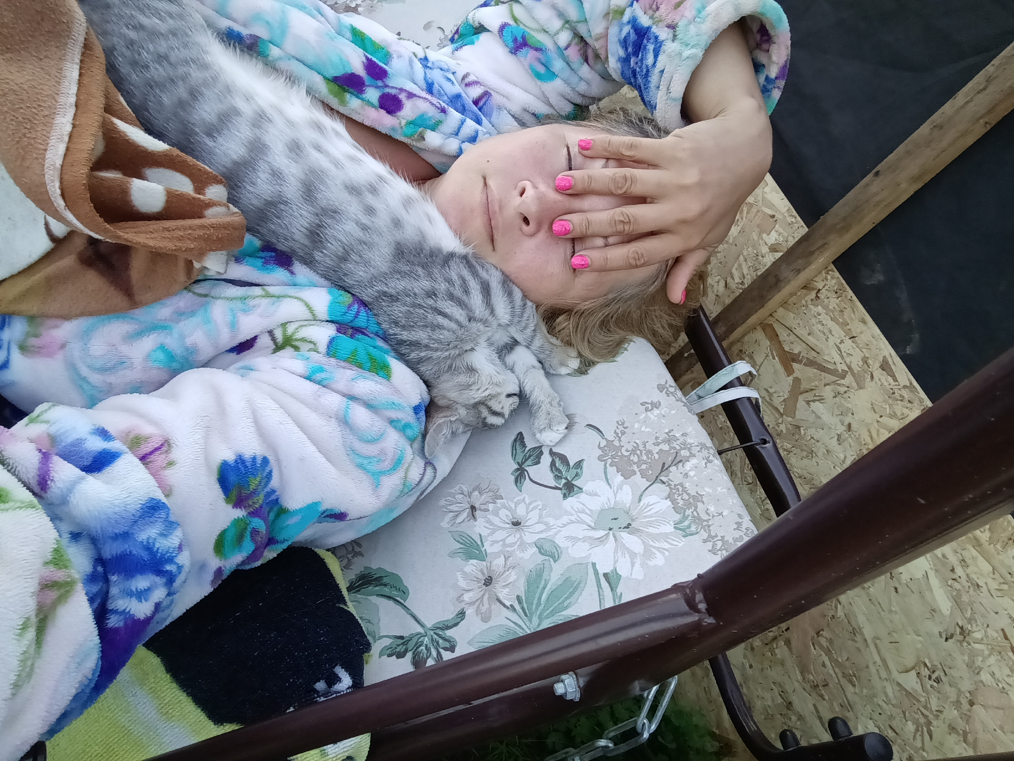 An overhead shot shows a woman lying down on her back with her eyes closed, covering them with her right hand. She is wearing a floral-patterned robe and has bright pink fingernails. A grey tabby cat is lying across her chest and neck, with its head resting near her chin. The woman is on a cushion with a floral pattern, which appears to be part of a swing or outdoor seating. In the background, there is a wooden surface and black fabric. The scene is outdoors, likely in a backyard or garden setting. The location in Ivanovskoe, Russia is not discernible from the image.