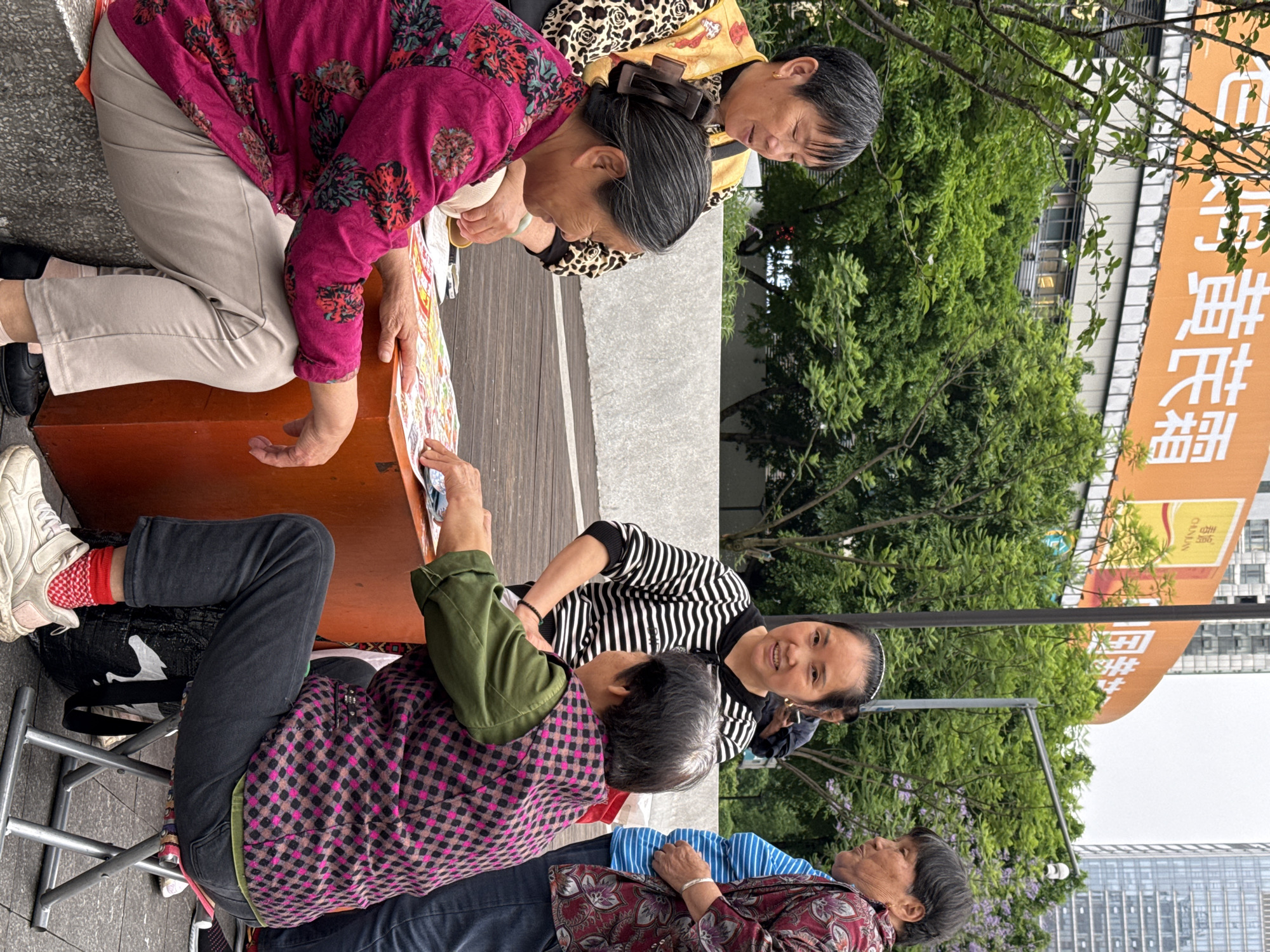 In Nan'an District, China, a group of elderly women is gathered around a low, orange table on an outdoor, paved surface. One woman in a pink floral top and beige pants kneels, looking at a colorful, possibly laminated, document on the table. Another woman in a leopard print top and yellow shirt stands and peers at the document over the kneeling woman's shoulder. Seated on a metal folding chair, a woman in a pink and black plaid shirt and green jacket massages the neck of another woman in a black and white striped shirt. Seated and partially visible is another woman in a floral top and striped pillow under her head. A large orange sign with white text is visible in the background with trees obscuring some of the sign. Tall buildings are visible in the background.