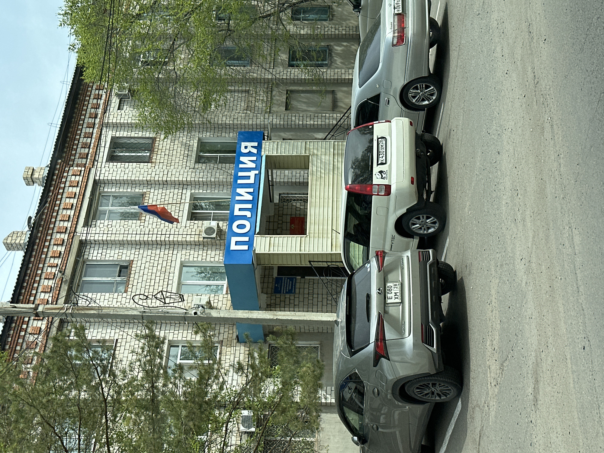 The image depicts the Police station in Raychikhinsk, Russia. The building is made of white brick and has a blue sign with the word "ПОЛИЦИЯ" (Police) written vertically in Russian. A Russian flag is mounted on the building. Three cars are parked in front of the station. The first car is a gray Lexus with the license plate "E 080 XM 28". The second car is a white Honda with the license plate "K 430 XM 28". The third car is a silver Toyota with the license plate "B 069 KX 28". Trees are present on either side of the building. Overhead power lines run across the top of the building. The sky is overcast.