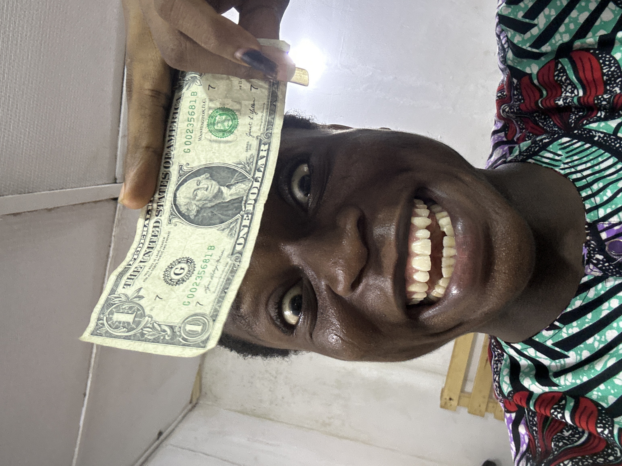 A close-up, tilted shot shows a smiling, dark-skinned woman holding a one-dollar bill over her forehead. The bill is slightly wrinkled and obscures the top of her head. Part of her patterned, brightly colored garment is visible on the right. The background is a plain, light-colored wall. Her teeth are prominently displayed in her wide smile, and her dark eyes are visible beneath the dollar bill. The specific location in the city of Unknown, Unknown is not discernible.