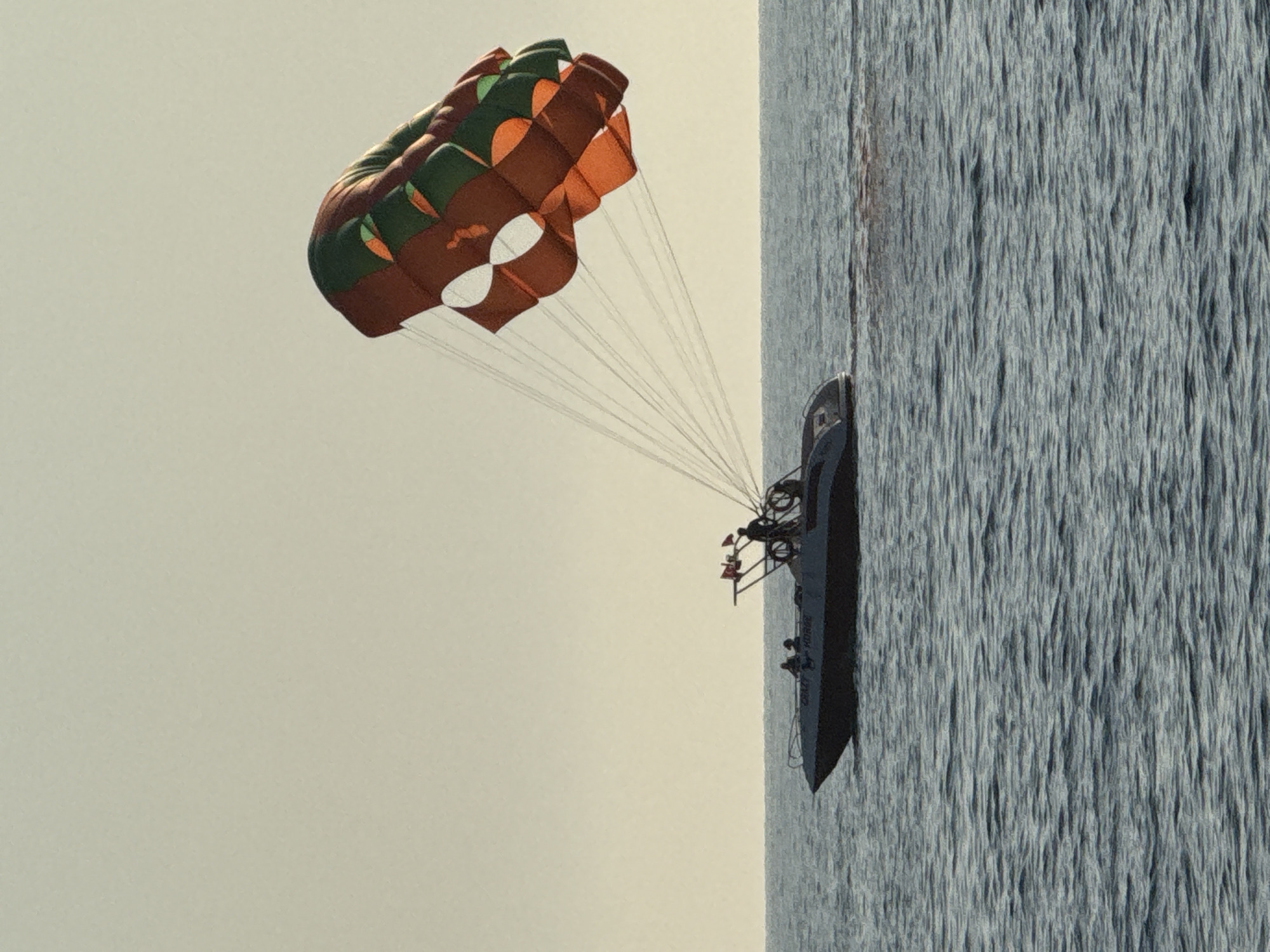 The image captures a parasailing activity in the waters off the coast of Alanya, Turkey. A speed boat, identified by the name "CRAZY HORSE" visible on its side, is moving through the choppy blue sea. Several individuals are visible on the boat, including two passengers seated and at least one person operating the boat. Above and behind the boat, a large, orange and green parachute is inflated and aloft, tethered to the boat by multiple lines. The parachute is partially obscured by the angle and the lighting. The sky above is a hazy, pale yellow, suggesting it may be late afternoon or early morning. The horizon is clearly defined between the sea and the sky.