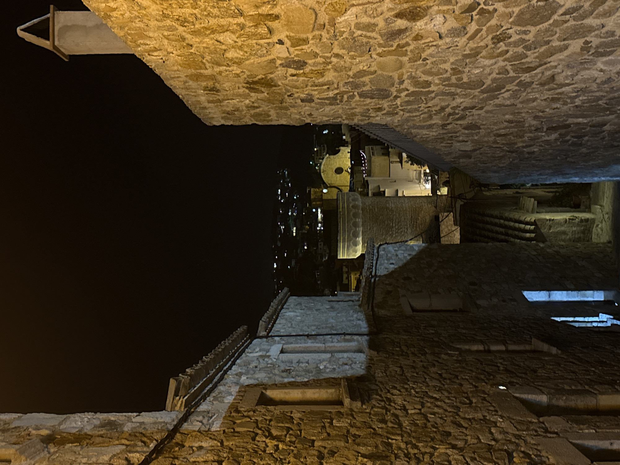 The photograph captures a nighttime view of a narrow, stone-walled alleyway in Tossa de Mar, Spain. The ancient stone buildings lining the alley are illuminated by artificial light, casting warm yellow and cool blue tones on their textured surfaces. Above, the night sky is a deep black.
In the distance, a cluster of buildings with illuminated windows creates a soft glow, indicating the presence of a town or city. A prominent church with a circular window and a steeple is visible amongst the distant structures, suggesting this alleyway is located within the historic center of Tossa de Mar, likely near the Vila Vella (old town). The architecture suggests a medieval or older settlement, characterized by its robust stone construction. A modern chimney with a triangular cap is visible atop one of the stone buildings in the foreground, contrasting with the aged materials.
The alley appears to be a pedestrian walkway, and there are no visible people or vehicles in the frame. The overall impression is one of historical depth and a quiet, nocturnal atmosphere within a Mediterranean coastal town. The materials and style of the buildings strongly suggest the distinctive architecture found in Tossa de Mar's old town, known for its well-preserved medieval fortifications and buildings.