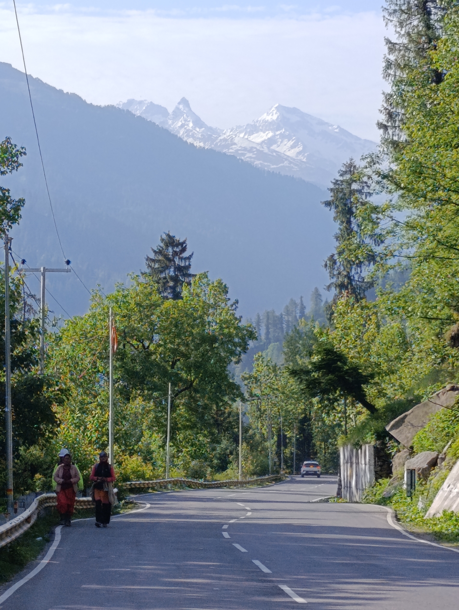 The image depicts a scenic mountain road in Solang, India. In the foreground, two individuals, dressed in traditional attire, are walking on the left side of the paved road. The road curves to the right, with a white railing bordering the left side. Lush green trees line the right side of the road, with some rocks and foliage visible on the hillside. In the distance, a white car is seen driving away from the camera. Further in the background, majestic snow-capped mountains rise against a hazy blue sky, indicative of a high-altitude region. Power lines and utility poles are present on the left side of the road, suggesting infrastructure in the area. The overall scene captures the natural beauty of the Solang valley.
