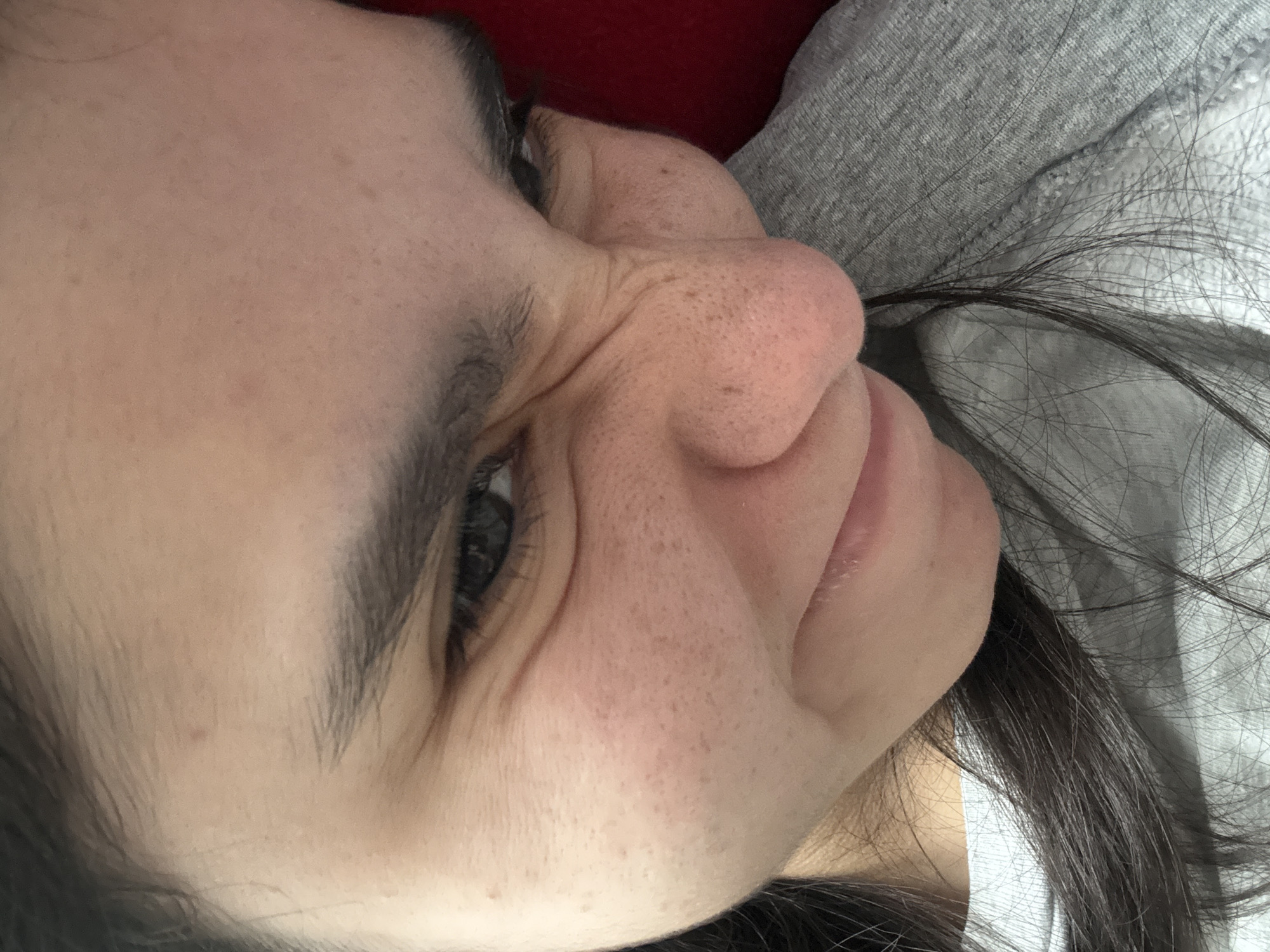 A close-up, indoor photograph shows a person's face, angled and slightly distorted due to the photographic perspective. The person has dark hair, which is partially visible around the face. One eye, with visible eyelashes and eyebrow, is open and looking towards the viewer's left. The skin on the face is fair, with visible pores and some slight freckling or pigmentation. The nose is prominent, and the mouth is partially visible. The person appears to be resting against a grey, possibly soft fabric, and a hint of red fabric is visible in the background. There are no identifiable landmarks or features to suggest the specific location within Austin, Texas, or any other location.
