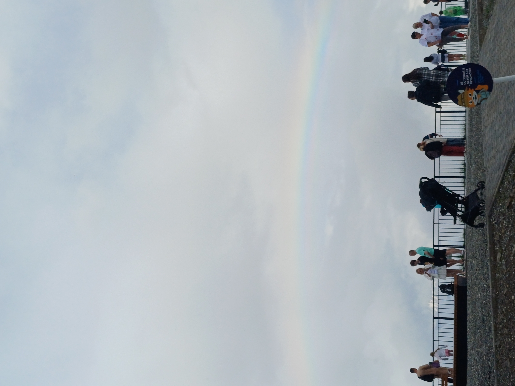 The image shows a group of people standing on an elevated walkway or observation deck, looking out at a cloudy sky with a faint rainbow. The walkway has a metal railing for safety. Below the walkway, a textured, pebbly surface is visible.
The subjects are diverse, including adults and children. Some are casually dressed, while others appear to be in athletic wear. They are standing in various poses, some looking at the rainbow, others conversing. There are also objects like strollers and bags present.
The scene appears to be an outdoor recreational area. The time of day is likely daytime, given the light, and the weather is overcast with a possibility of rain, as indicated by the rainbow. The location is identified as Estosadok, Russia.
A circular sign with text and a cartoon animal illustration is visible on a pole. The text on the sign appears to be in Cyrillic. While not fully legible, parts of it can be deciphered. The cartoon animal appears to be a fox or a similar creature.
The overall mood of the scene is relaxed and observational. The presence of the rainbow suggests a brief moment of beauty amidst the cloudy weather. The people seem to be enjoying the view and the atmosphere.